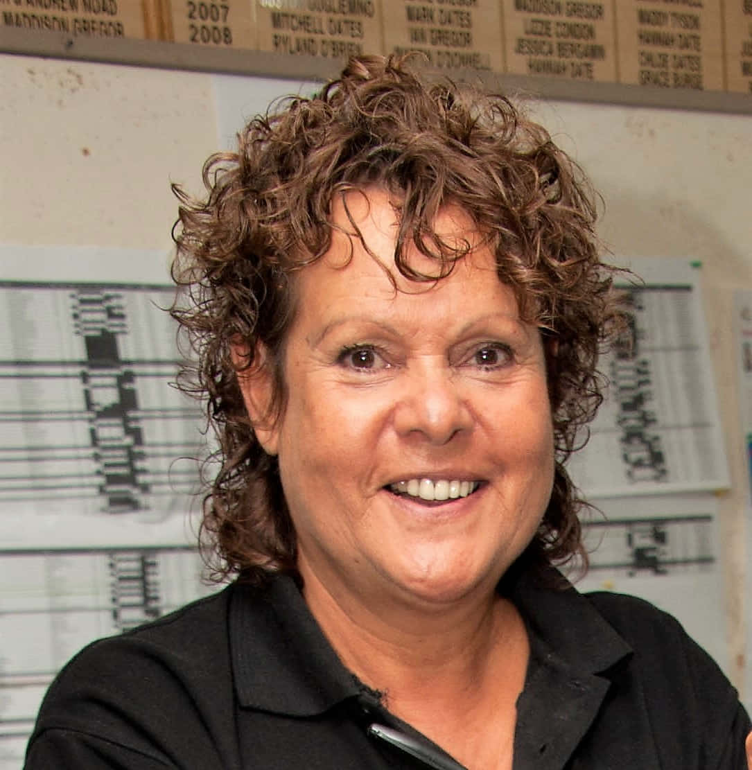 網球選手 Evonne Goolagong Cawley 頭像照 壁紙 — 一位微笑的女士，棕色捲髮，身穿黑色襯衫，背景是運動隊橫幅和圖表。