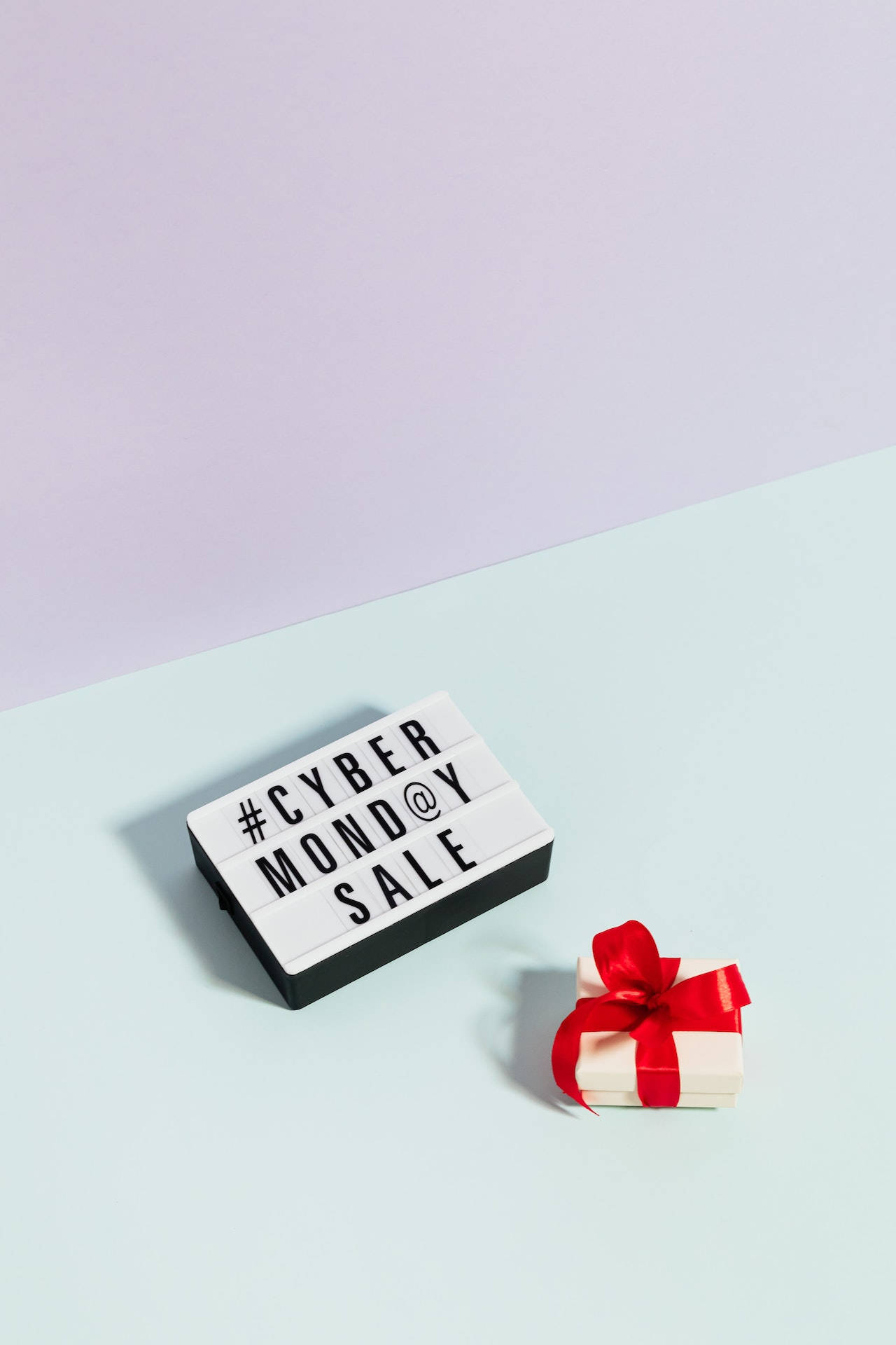 網路購物節禮盒 壁紙 — 極簡風格的平鋪構圖，一個燈箱顯示 #CYBERMONDAY SALE，旁邊是一個繫著紅色蝴蝶結的小白色禮物盒，背景為雙色粉彩。