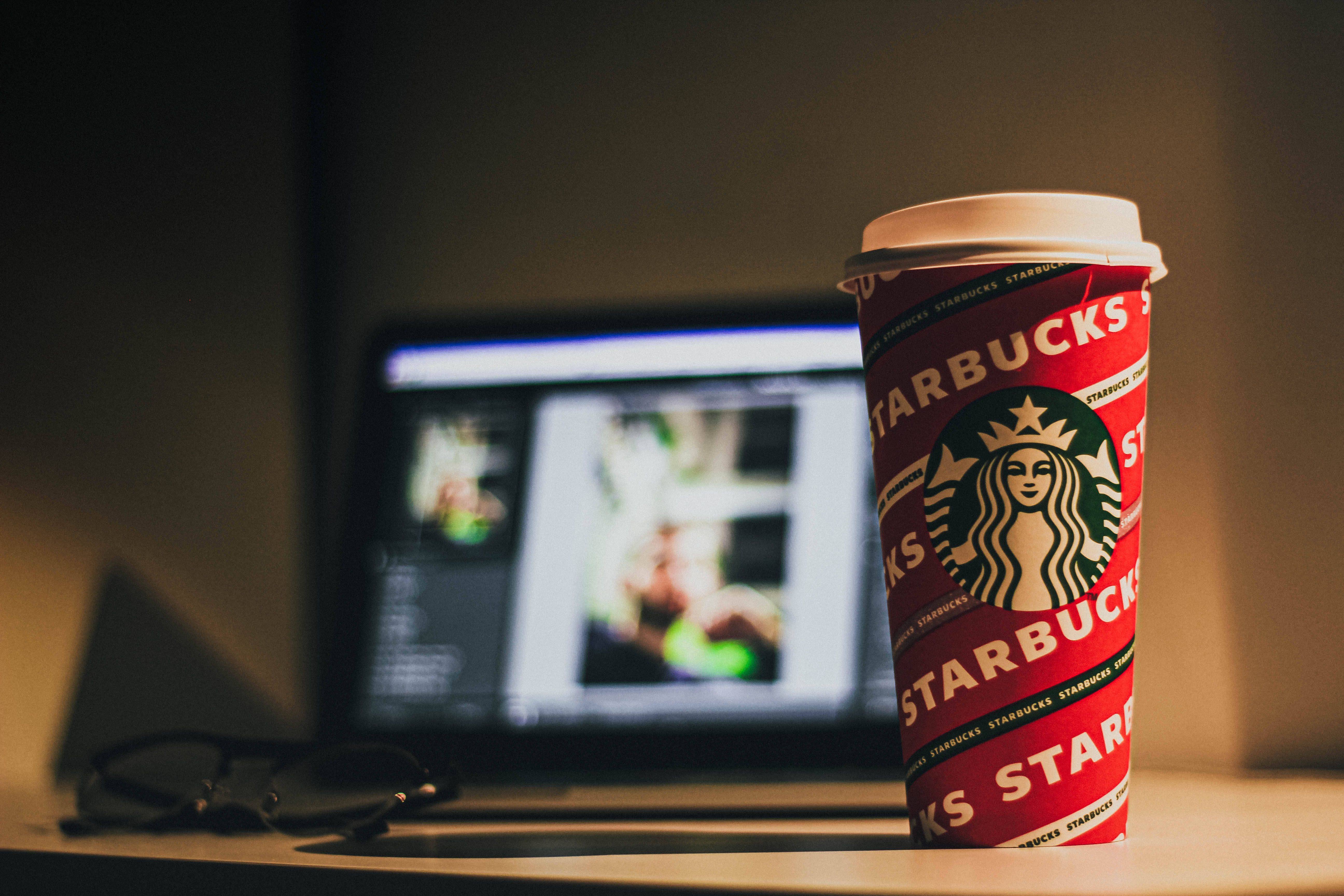 編輯軟體與星巴克杯 壁紙 — 一個紅色Starbucks咖啡杯放在溫暖光線下的桌上，旁邊有手提電腦和眼鏡。