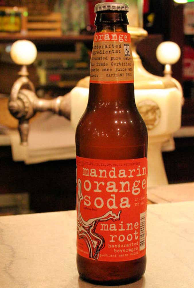 緬因州柑橘味汽水飲品 壁紙 — 一個琥珀色玻璃瓶裝的柑橘橙味汽水，帶有鮮明的紅色標籤，放在一個帶有復古水龍頭的酒吧櫃檯上，光線柔和溫暖。