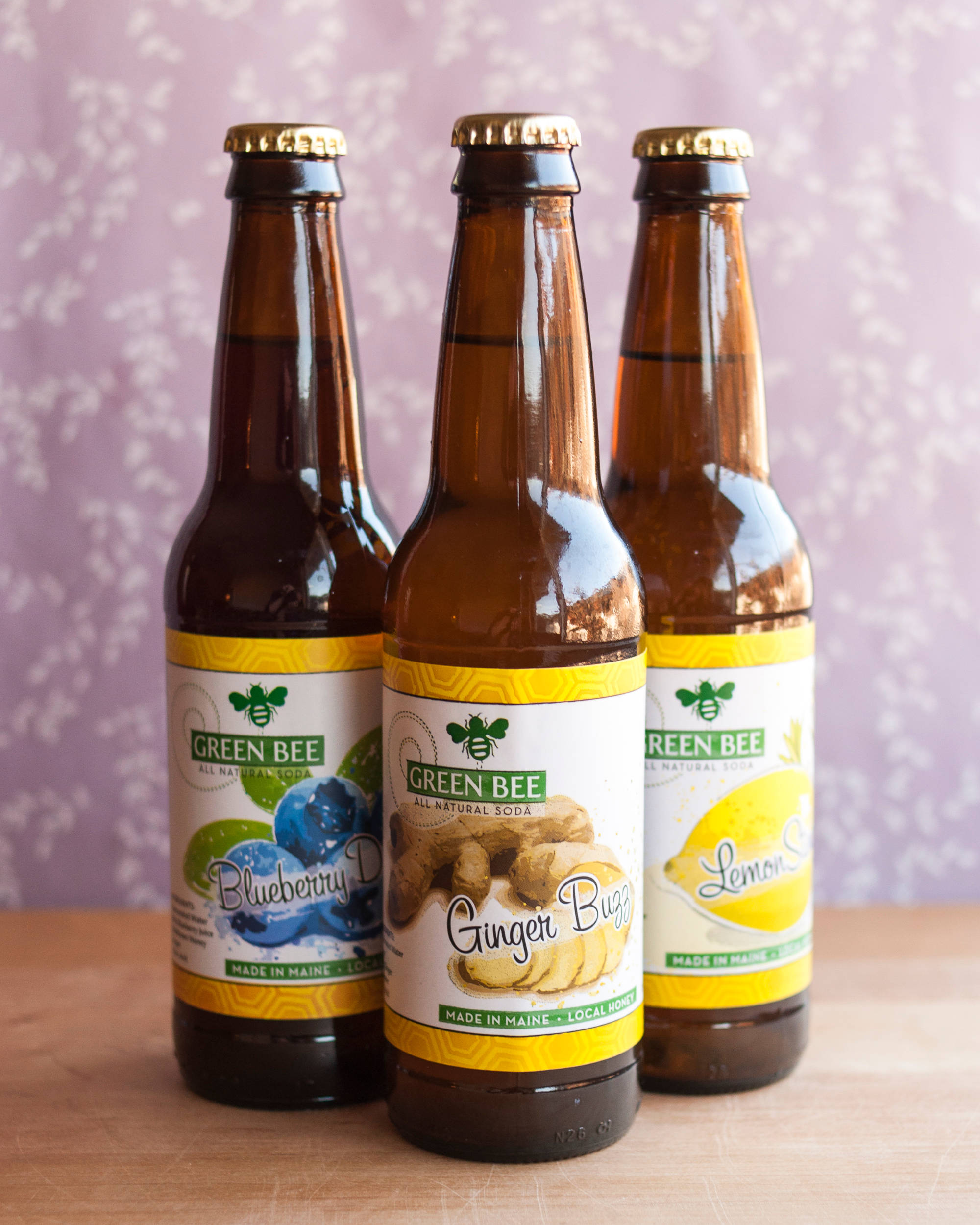 緬因州蘇打飲料三種口味 壁紙 — 三瓶琥珀色玻璃瓶的Green Bee全天然汽水，有藍莓薑蟲和檸檬口味，放在木質表面上，背景是柔和的紫色花卉。