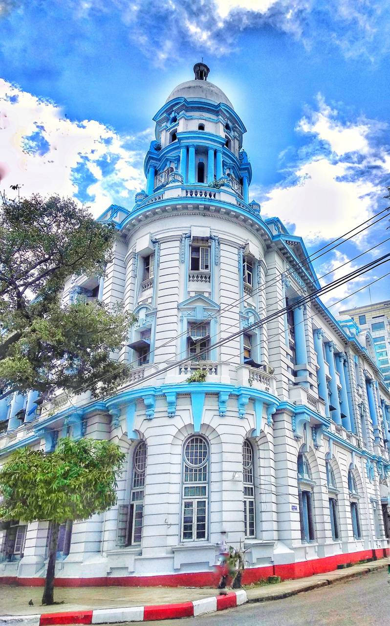 緬甸經濟銀行仰光市分行 壁紙 — 一座宏偉的新古典主義建築，帶有白色和充滿活力的藍色點綴，在藍天白雲下高高聳立。