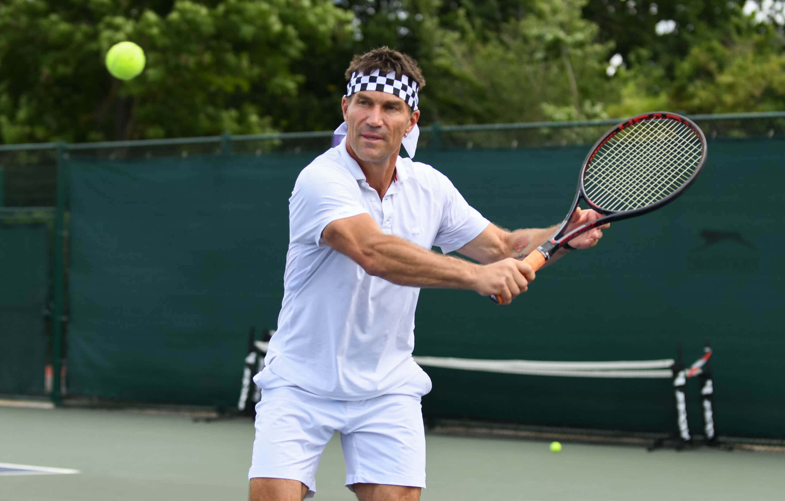 練習時光 Pat Cash 壁紙 — 一名身穿白色裝備的男性網球手在綠色的球場上揮拍擊打一個明亮的黃色網球，球場周圍有樹木。