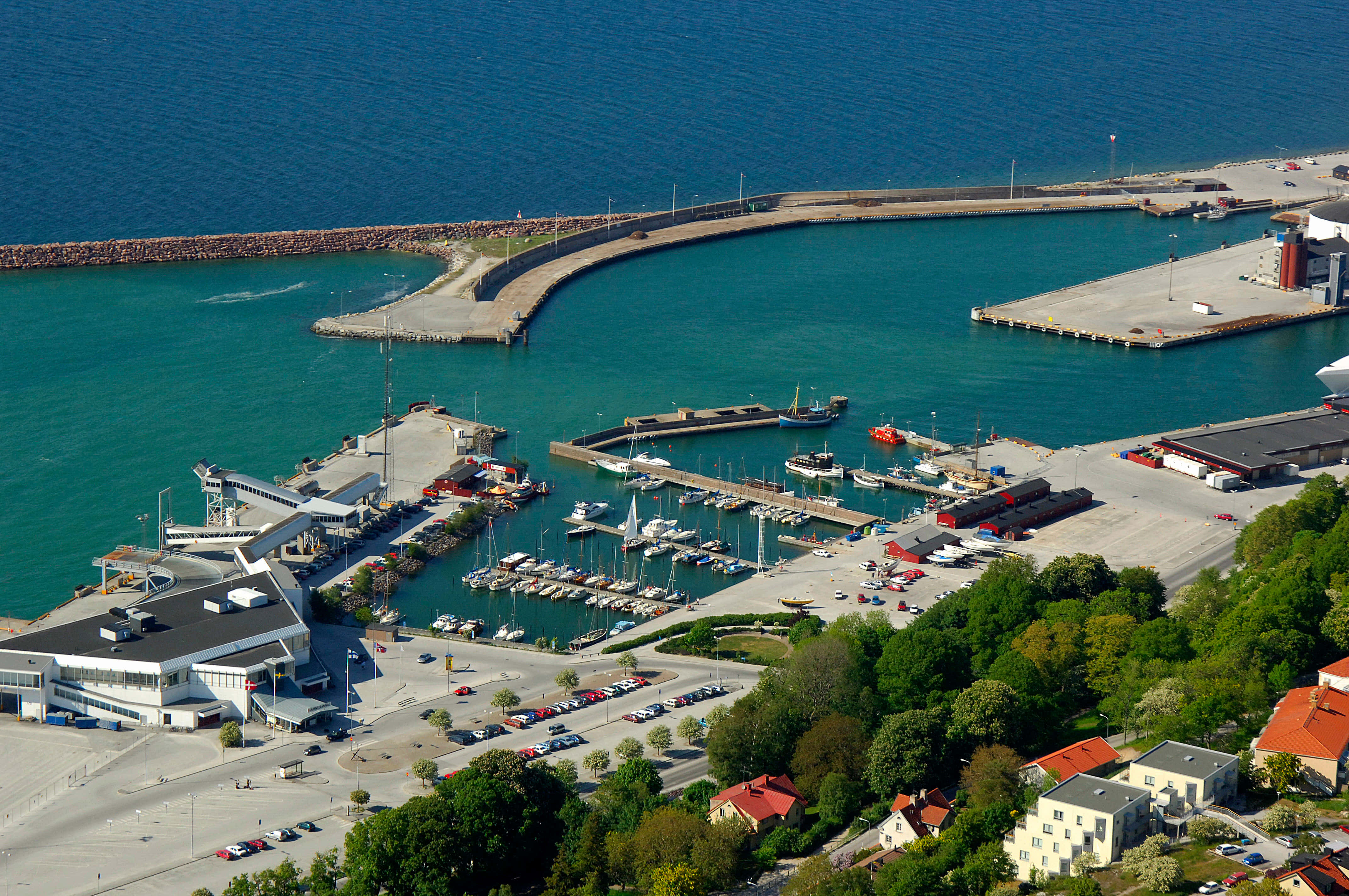 Visby 桌布 壁紙 — 繁忙港口的鳥瞰圖，有碧綠色的海水、遊艇碼頭、船隻、碼頭和周圍的綠樹，在明亮的日光下。