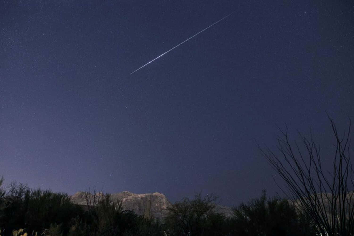 繁星滿天的夜空中，流星雨綻放絢麗色彩 壁紙 — 一顆明亮的流星劃過星空夜空，下方是剪影般的沙漠山脈和稀疏的植被。