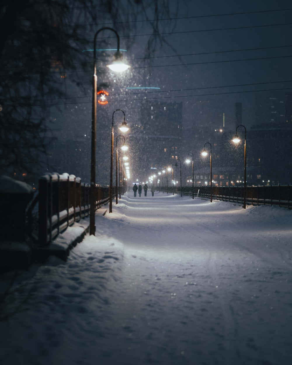 雪落在燈火通明的城市步道上，遠處有人行走。