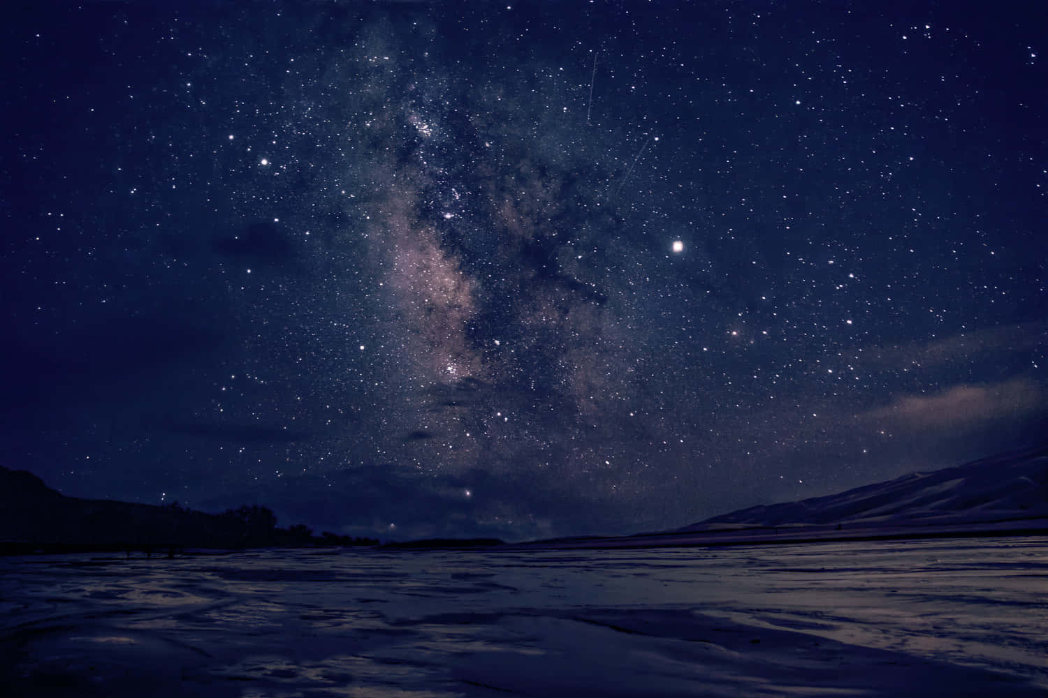 繁星滿天的夜空與沙丘 壁紙 — 令人嘆為觀止的冬夜天空，充滿無數星星和銀河，拱形在白雪覆蓋的風景和遠處的山脈之上。