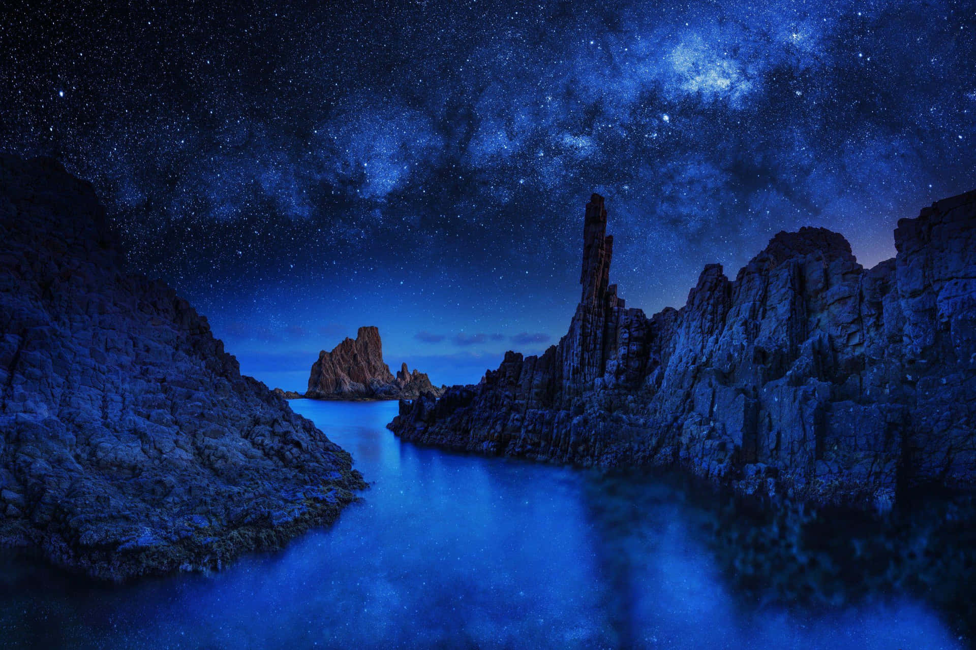 繁星點點的夜空，銀河在平靜的藍色海洋和岩石懸崖上空。