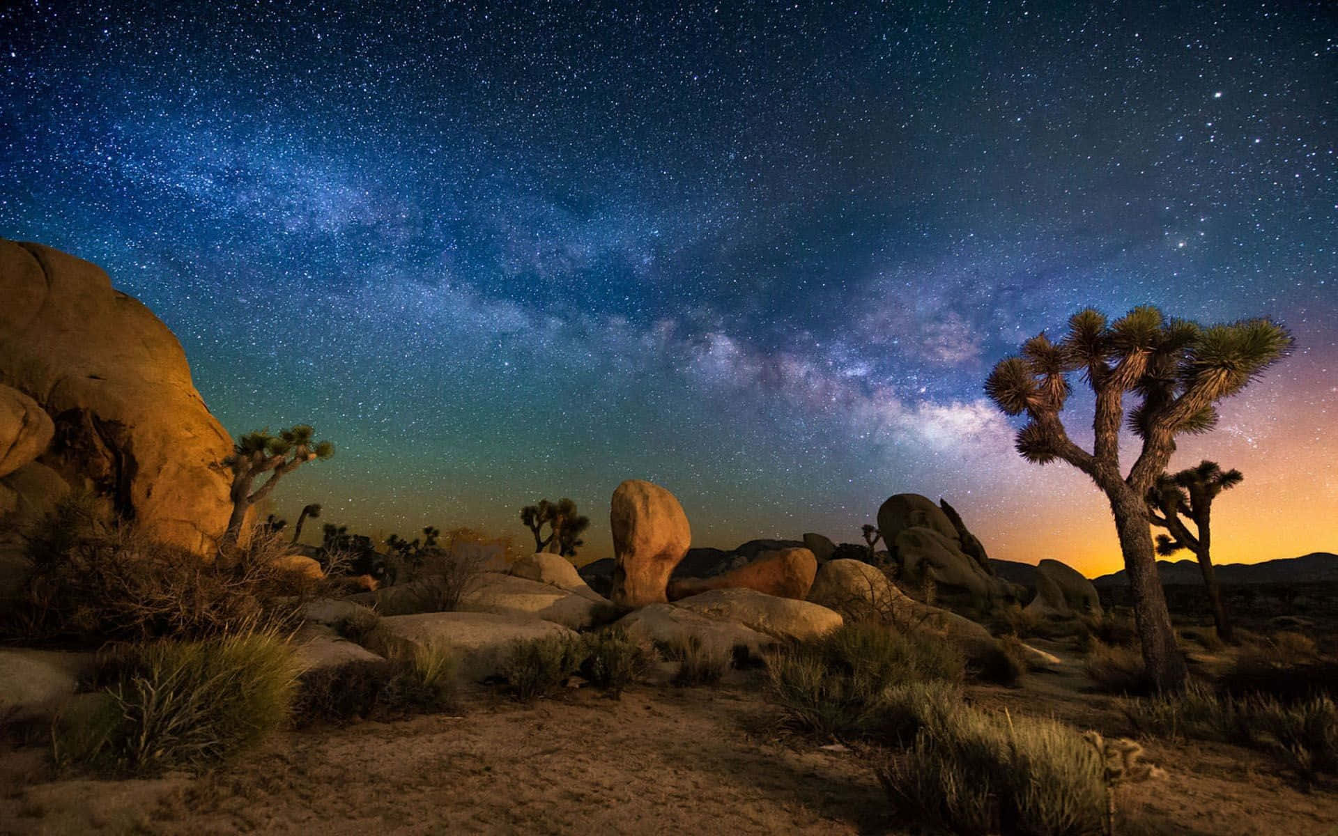繁星滿天約書亞樹圖 壁紙 — 繁星點點的夜空，銀河拱跨沙漠景觀，點綴著約書亞樹和巨石。