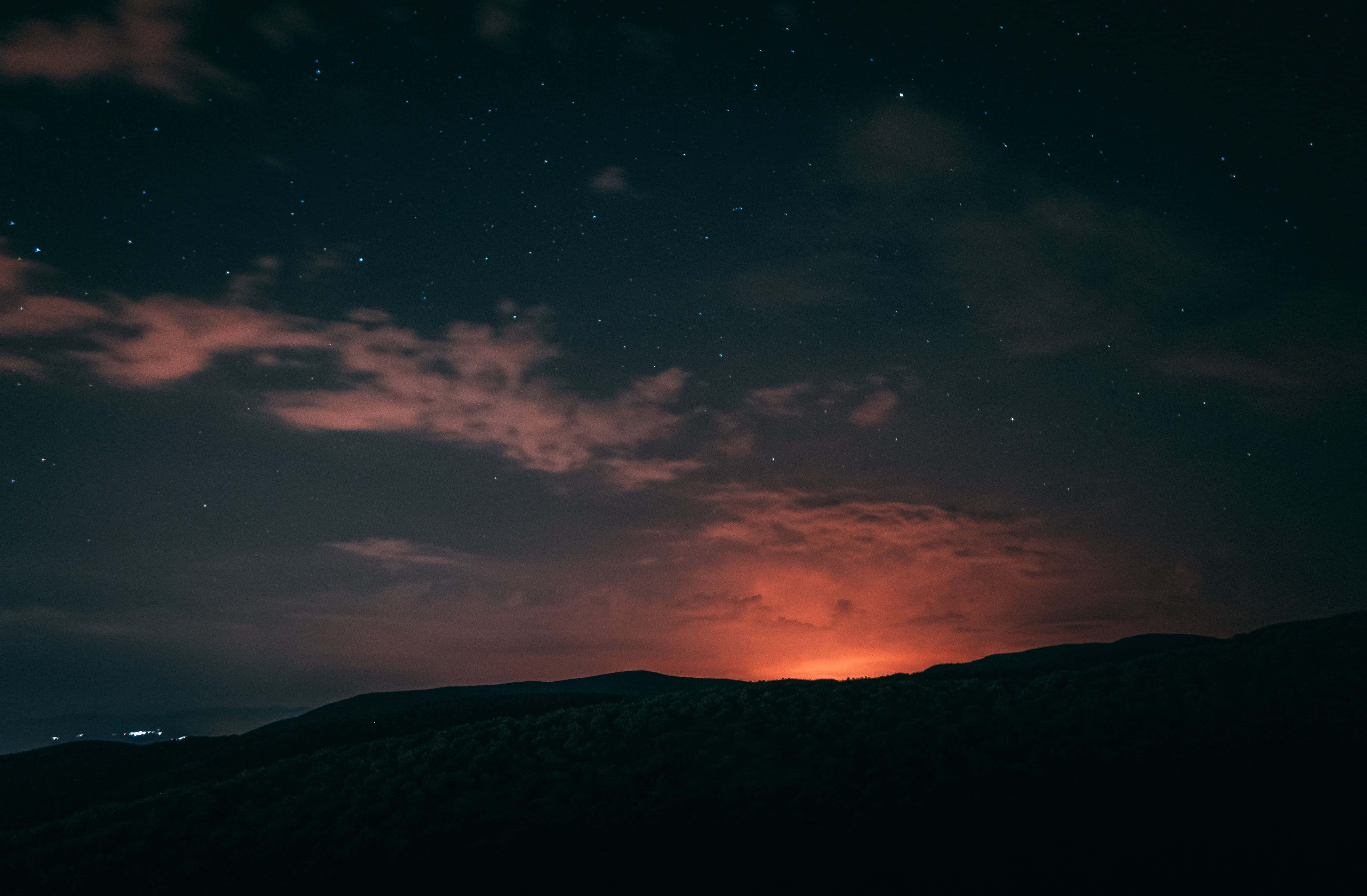 Horizon 桌布 壁紙 — 繁星點點的夜空，在黑暗的山脈輪廓和遠處的城市燈光之上，有發光的橙色雲彩。