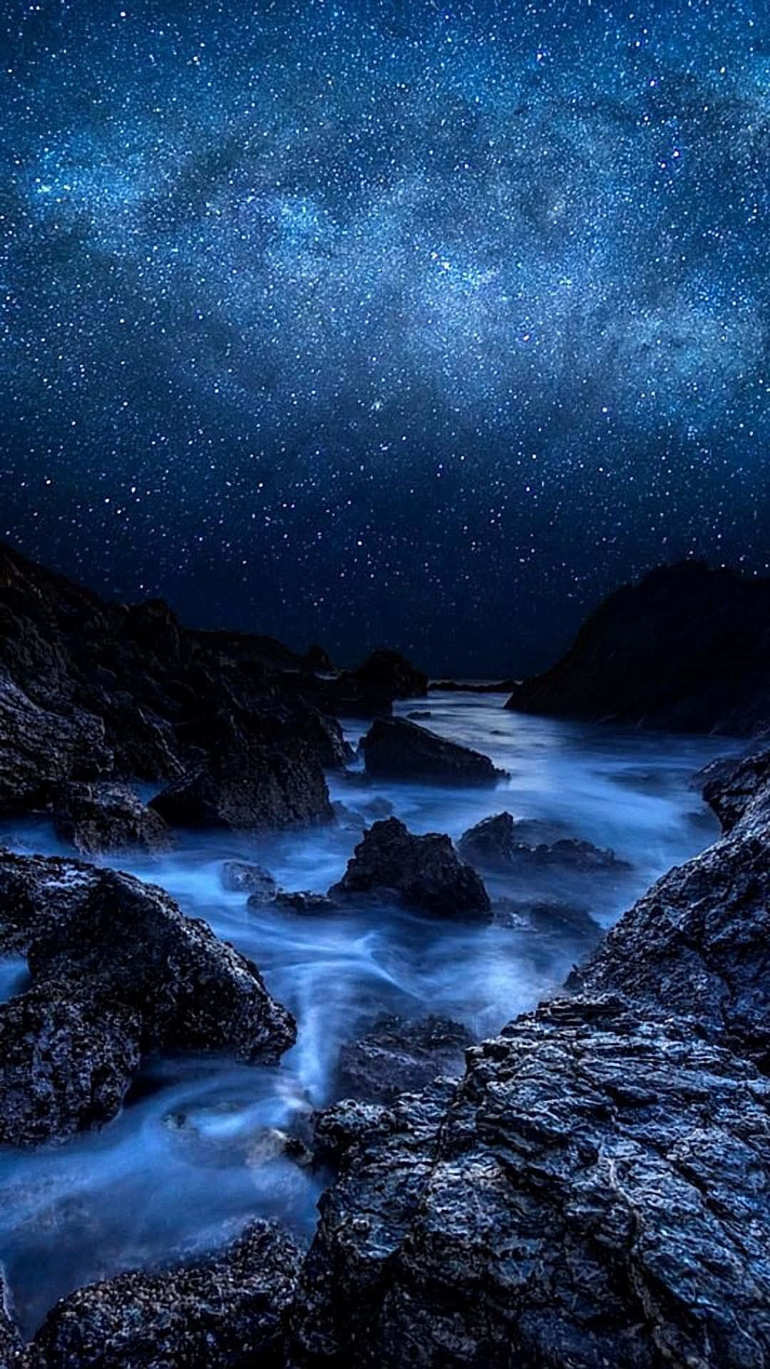 平板電腦 桌布 壁紙 — 繁星點點的夜空，銀河拱跨月光照耀的岩石海岸線，發光的藍色海水在深色石頭之間流動。