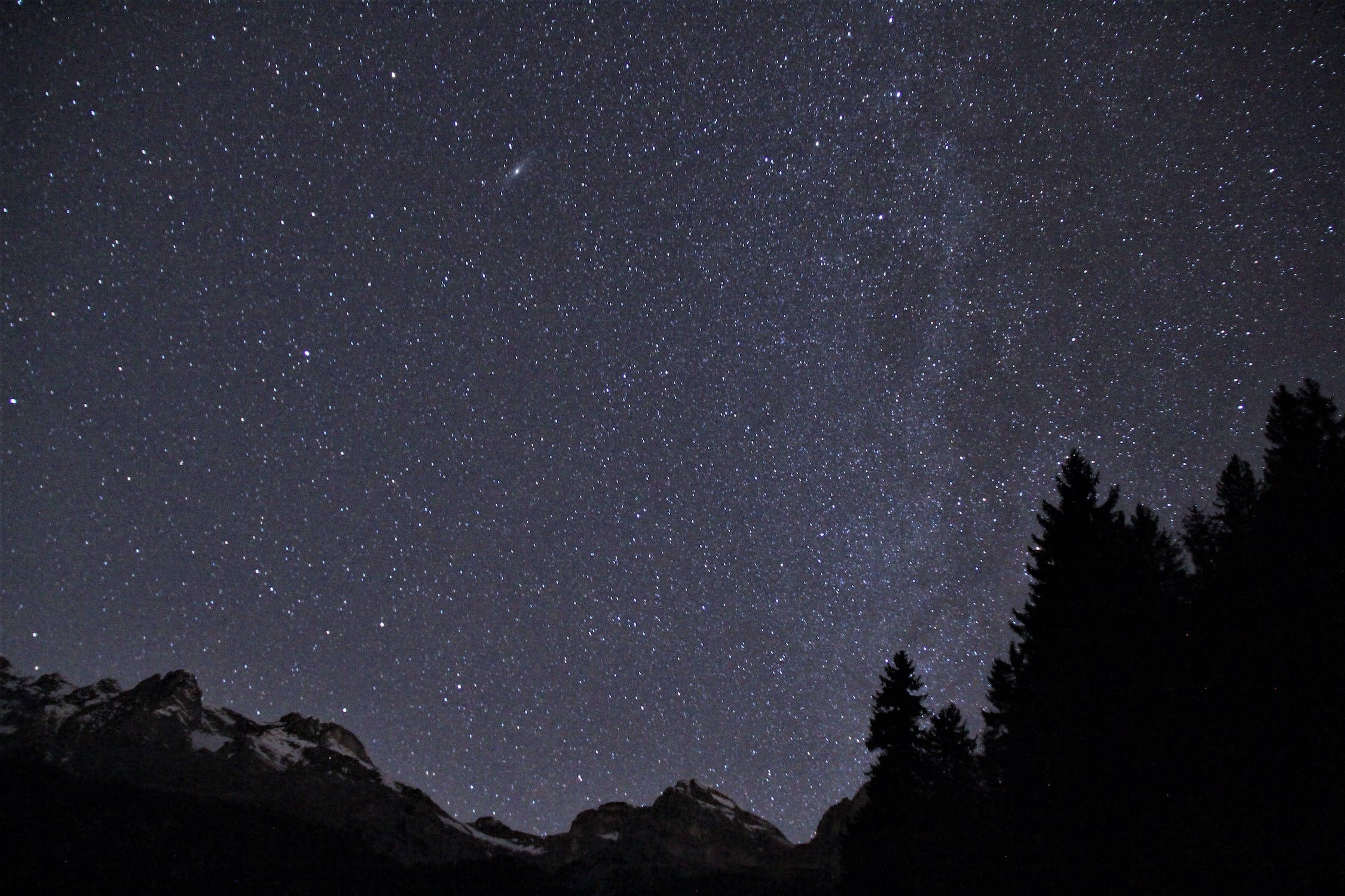 星星 桌布 壁紙 — 繁星點點的夜空拱起在積雪的山峰和漆黑的松樹之上，松樹在宇宙之光中形成剪影。