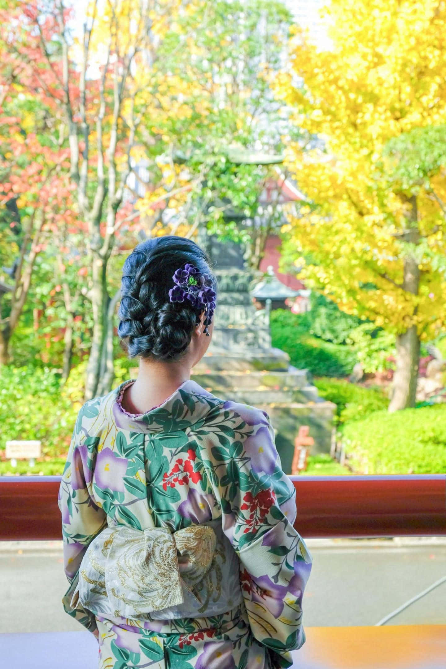 繽紛和服佈局 壁紙 — 一位穿著花卉和服的女士站在木製陽台上，凝視著被鮮豔秋樹環繞的寺廟。