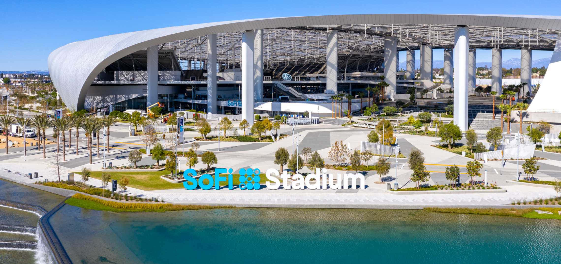 置身於NFL最大體育場SoFi Stadium的熱鬧中心 壁紙 — 現代SoFi Stadium的鳥瞰圖，在晴朗的藍天下，前景有棕櫚樹和藍綠色水景。
