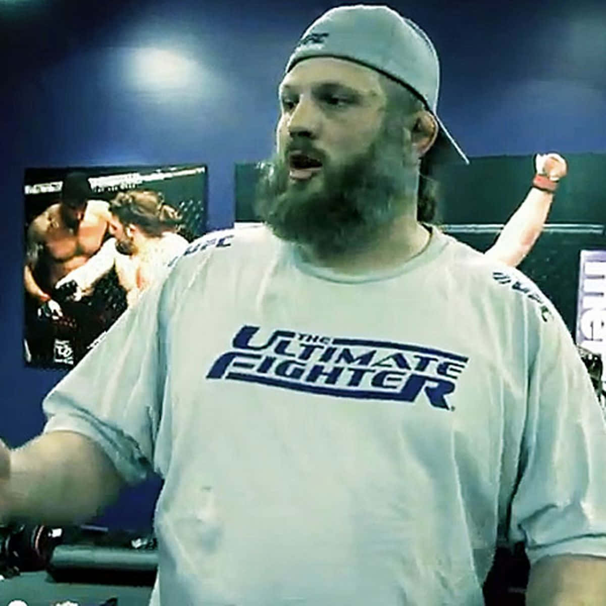 留鬍鬚的男子穿著UFC T恤在健身室中與搏擊海報一起做手勢。