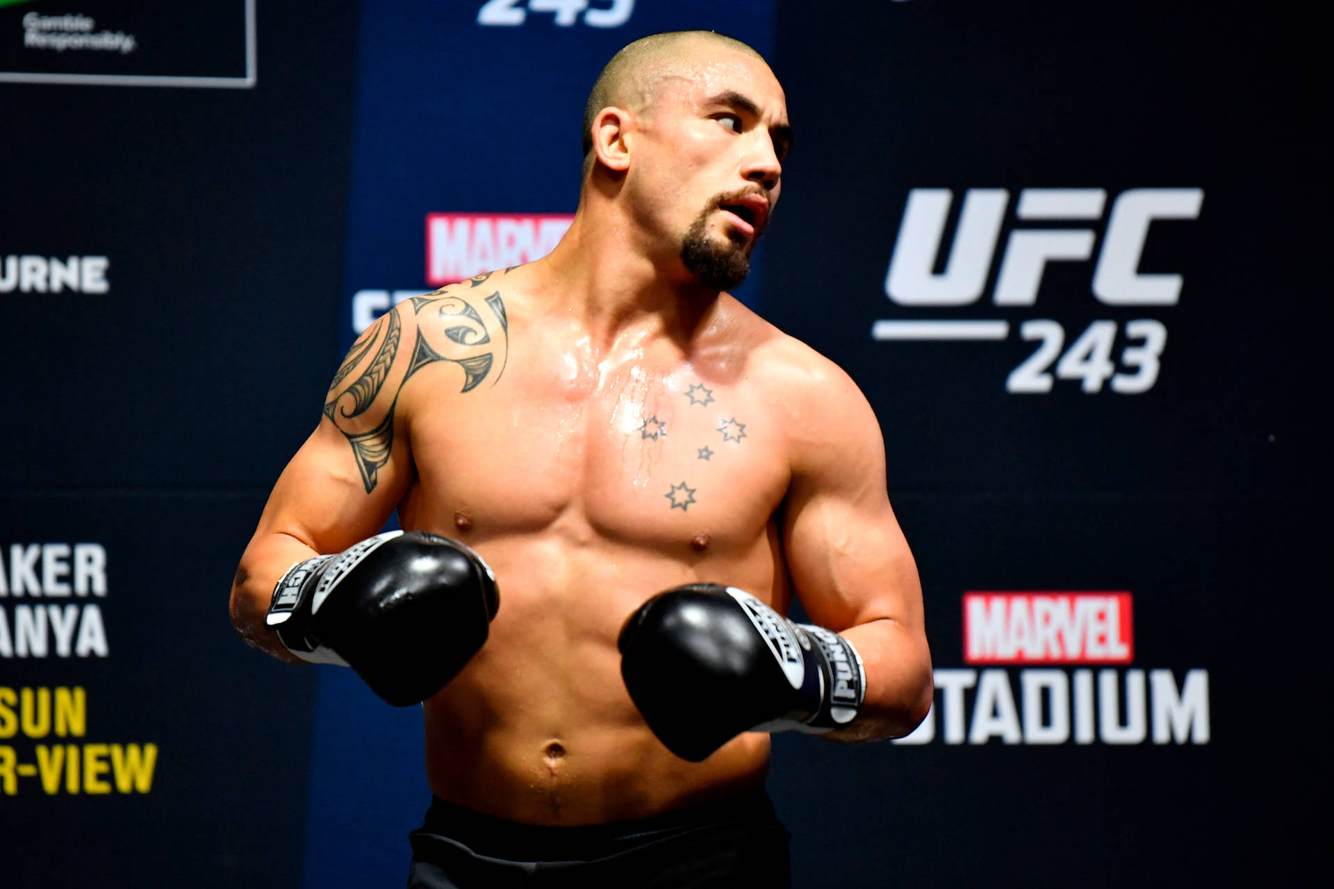 羅伯特·惠特克戴拳擊手套 壁紙 — 一位肌肉發達的UFC拳手，身上有紋身，戴著黑色手套，在藍色活動背景前充滿氣勢地站立。