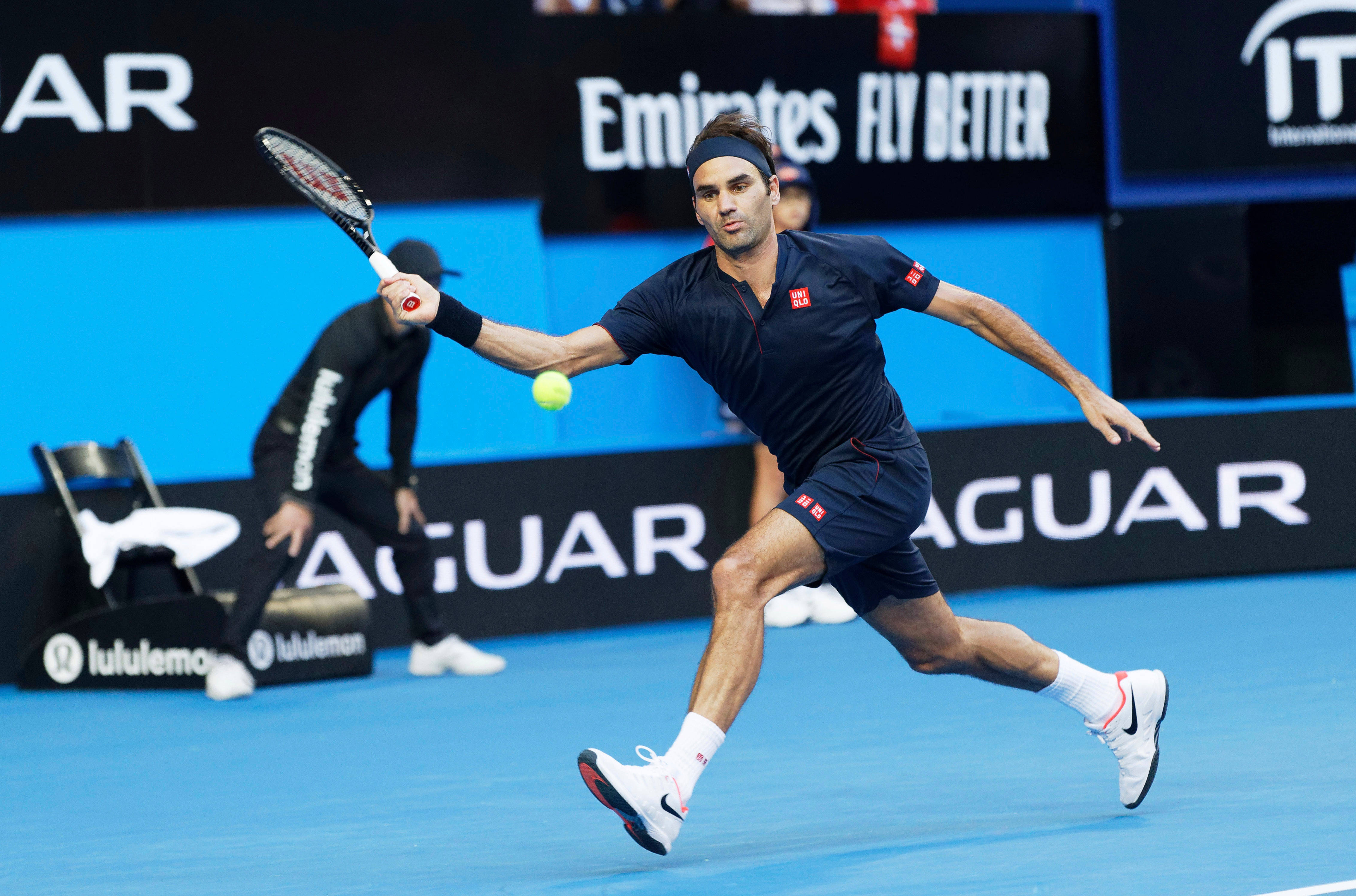 羅傑·費德勒藍色球衣 壁紙 — 網球手 Roger Federer 在揮拍中途向前猛衝，在明亮的體育場燈光下於充滿活力的藍色球場上擊打黃色球。