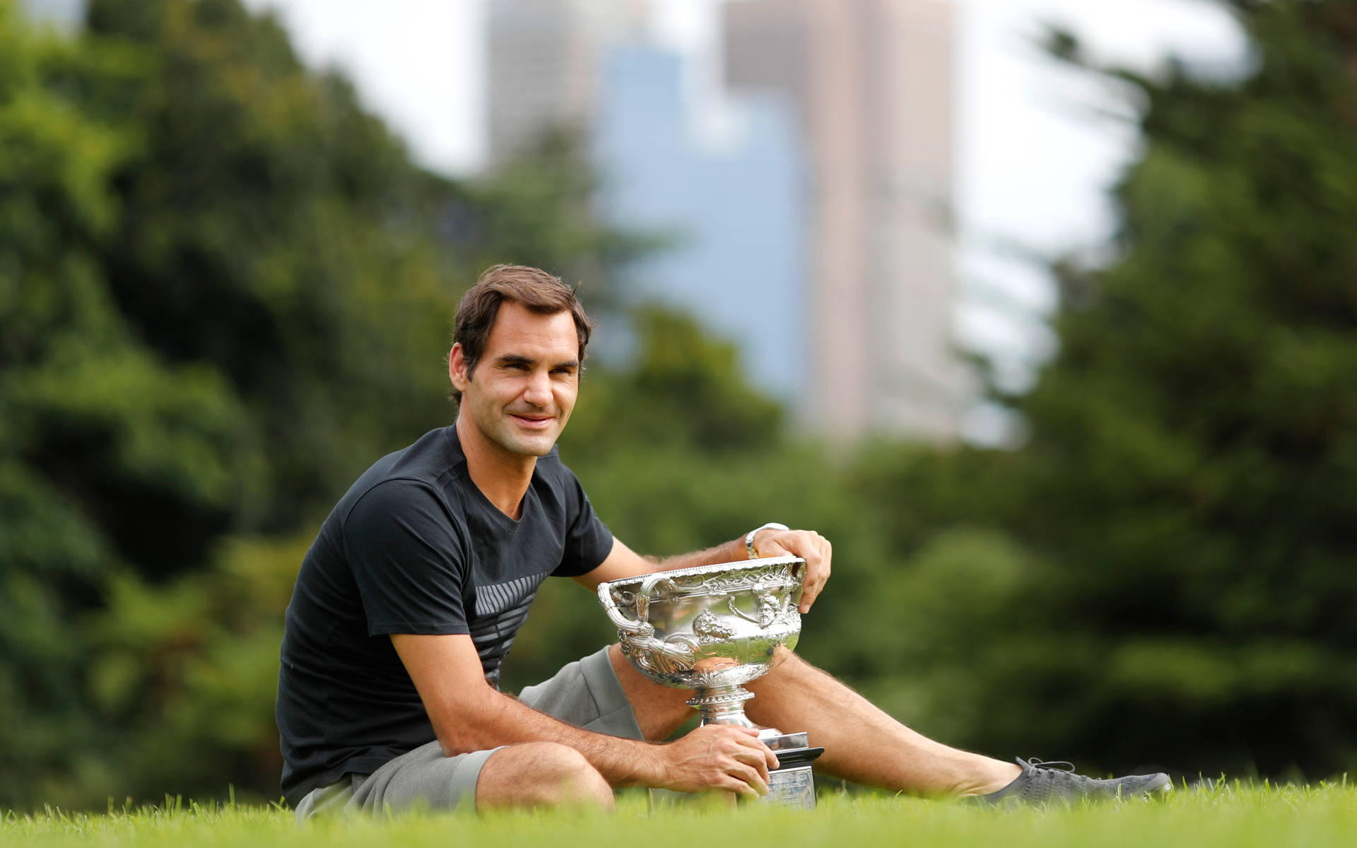 羅傑·費德勒高舉澳洲網球公開賽男子冠軍獎盃 壁紙 — Roger Federer 坐在草地上，手持銀色獎盃，背後的城市摩天大樓模糊不清。