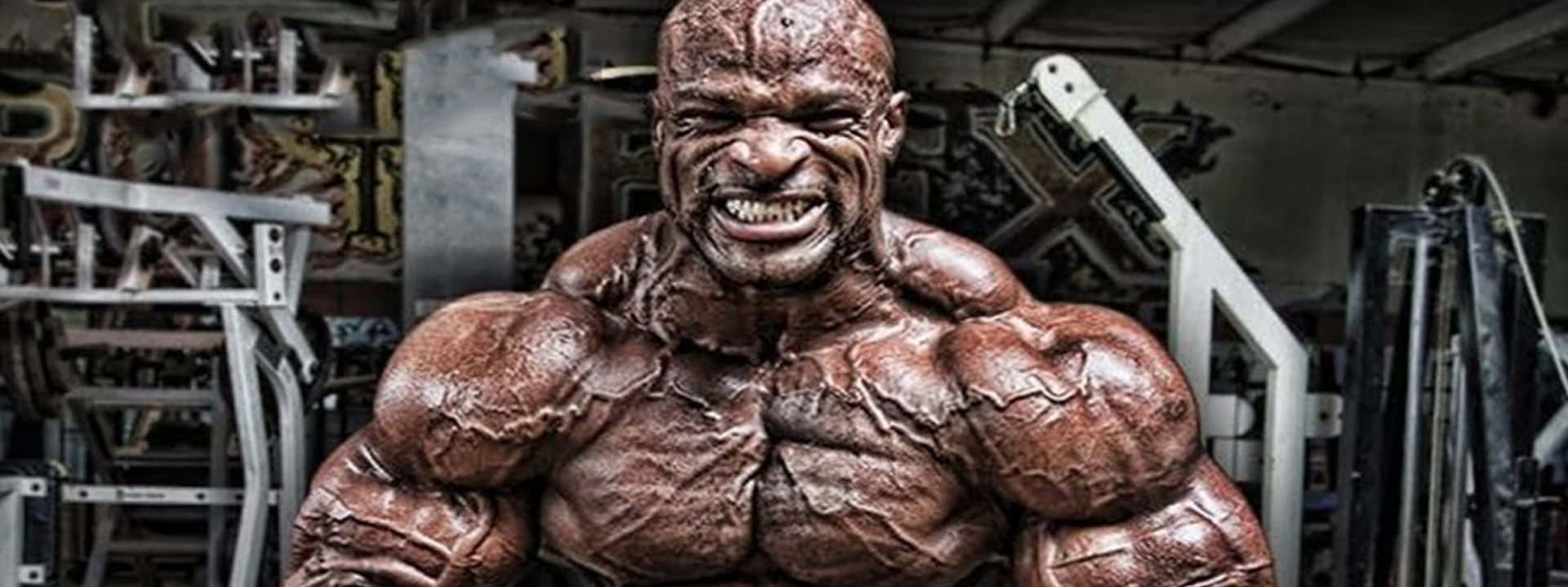 羅尼·柯爾曼肌肉血管明顯 壁紙 — 一個肌肉發達、露出兇猛笑容的男人站在一個雜亂的健身房裡，周圍都是舉重器材。