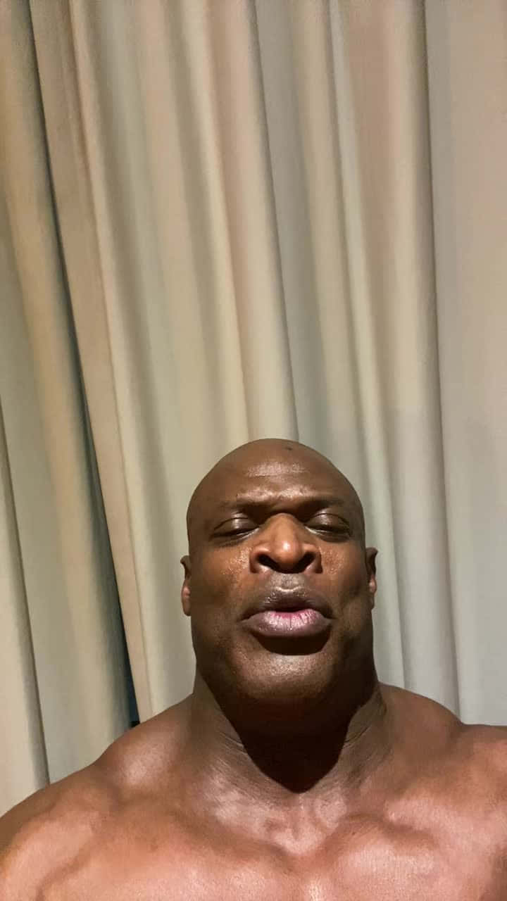一個赤裸上身的肌肉發達光頭男人閉著眼睛，在室內靠近米色窗簾站立。