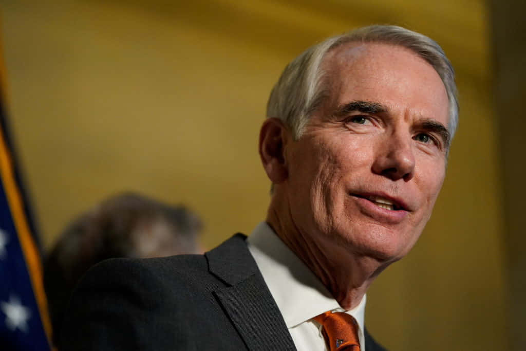 羅布·波特曼對著天空的肖像 壁紙 — 一名身穿西裝、打橙色領帶的男子近距離說話，背景是模糊的美國國旗。