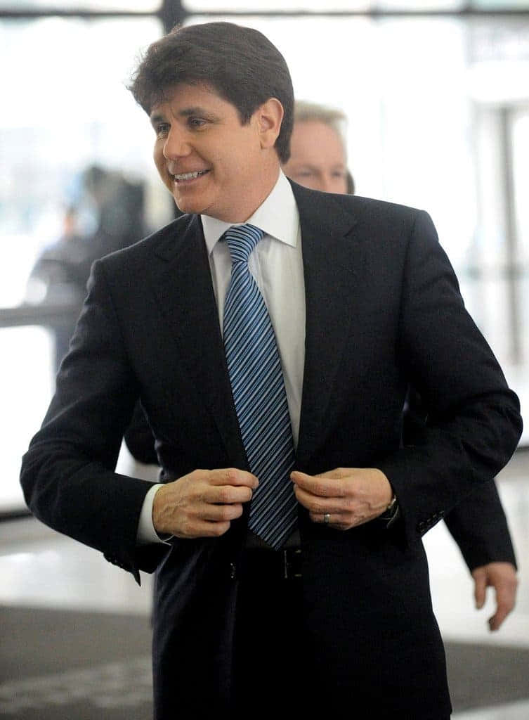 羅德·布拉戈耶維奇整理外套鈕扣 壁紙 — 一位微笑的男子身穿深色西裝和條紋領帶，在明亮的室內環境中扣上外套的鈕扣。