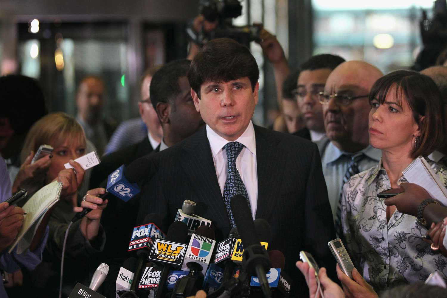 羅德·布拉戈耶維奇被記者與麥克風包圍 壁紙 — 一名身穿西裝的男子在擠滿人的記者室中，被麥克風和攝影機包圍，向記者發言。