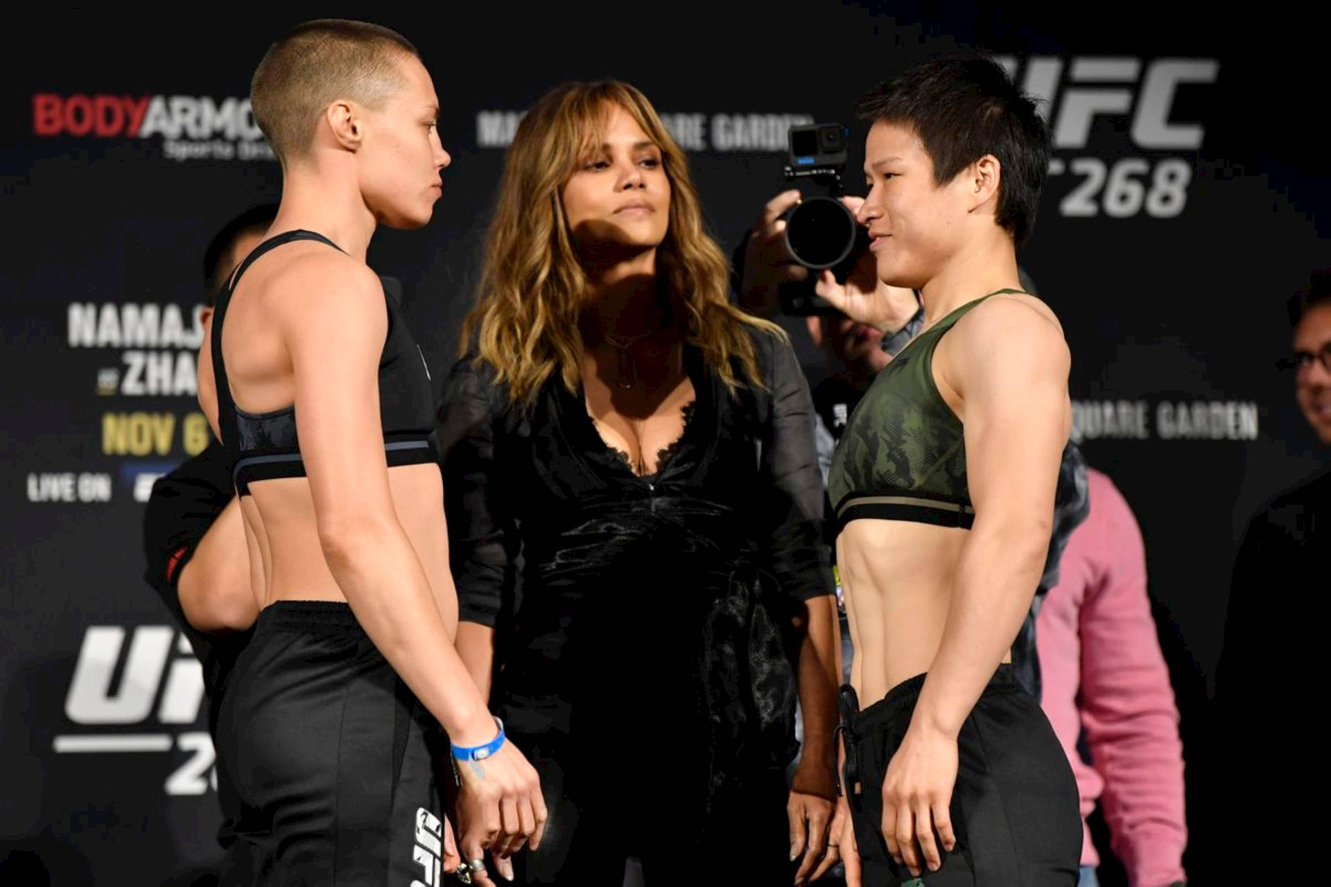 羅斯·納馬朱斯、哈莉·貝瑞與張偉麗 壁紙 — 兩位UFC女拳手在稱重活動中對峙，中間有一位名人。