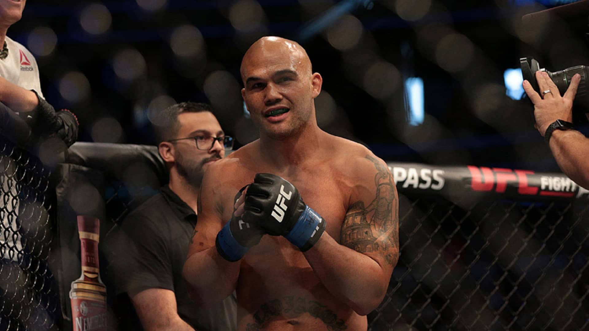 羅比·勞勒在UFC賽事中慶祝勝利 壁紙 — 一位帶有紋身的勝利UFC拳手在八角籠內微笑，背景是明亮的燈光和模糊的人群。