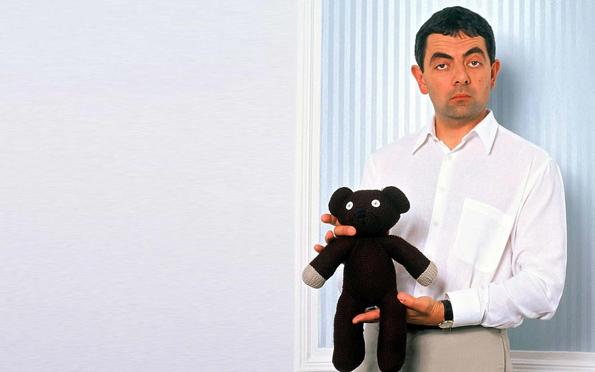 Mr Bean 桌布 壁紙 — 羅溫·艾金森飾演戇豆先生，手持一隻細小的棕色編織泰迪熊，背景是條紋牆壁，並有空白白色空間。