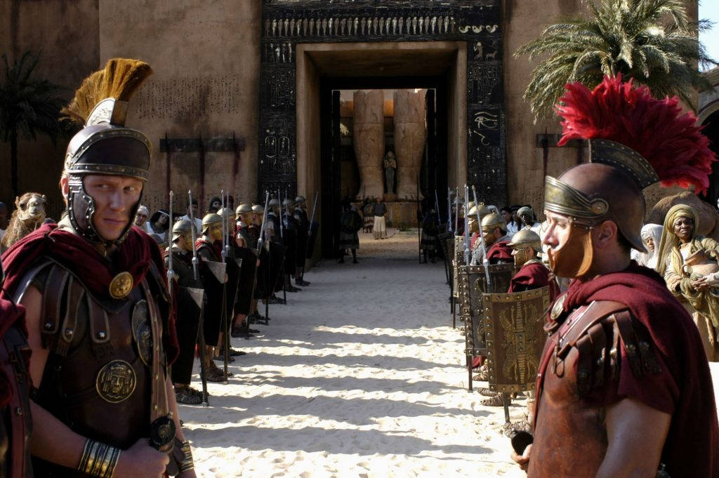 羅馬2：羅馬角鬥士與將領 壁紙 — 身穿精細盔甲的羅馬軍團士兵在陽光明媚的沙漠大門前立正，兩旁有棕櫚樹和象形文字牆。