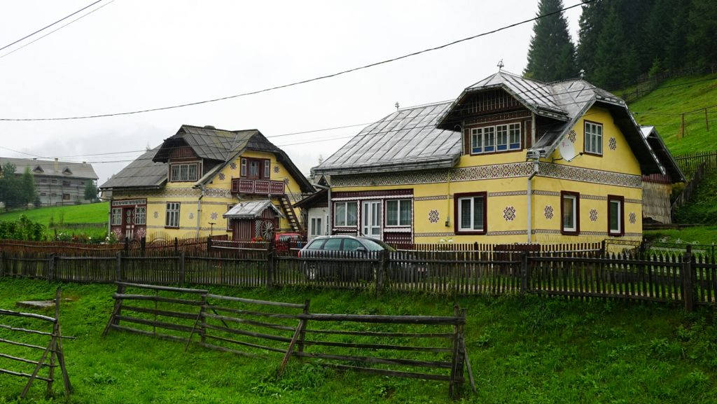 羅馬尼亞 Ciocănești 村莊風景 壁紙 — 傳統黃色木屋帶有裝飾圖案，坐落在陰天下的綠色山坡上。
