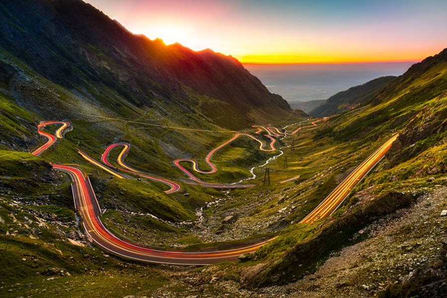羅馬尼亞布蘭城堡迷人風景 壁紙 — 日落時分蜿蜒的山路，發光的汽車軌跡蜿蜒穿過翠綠的山谷。