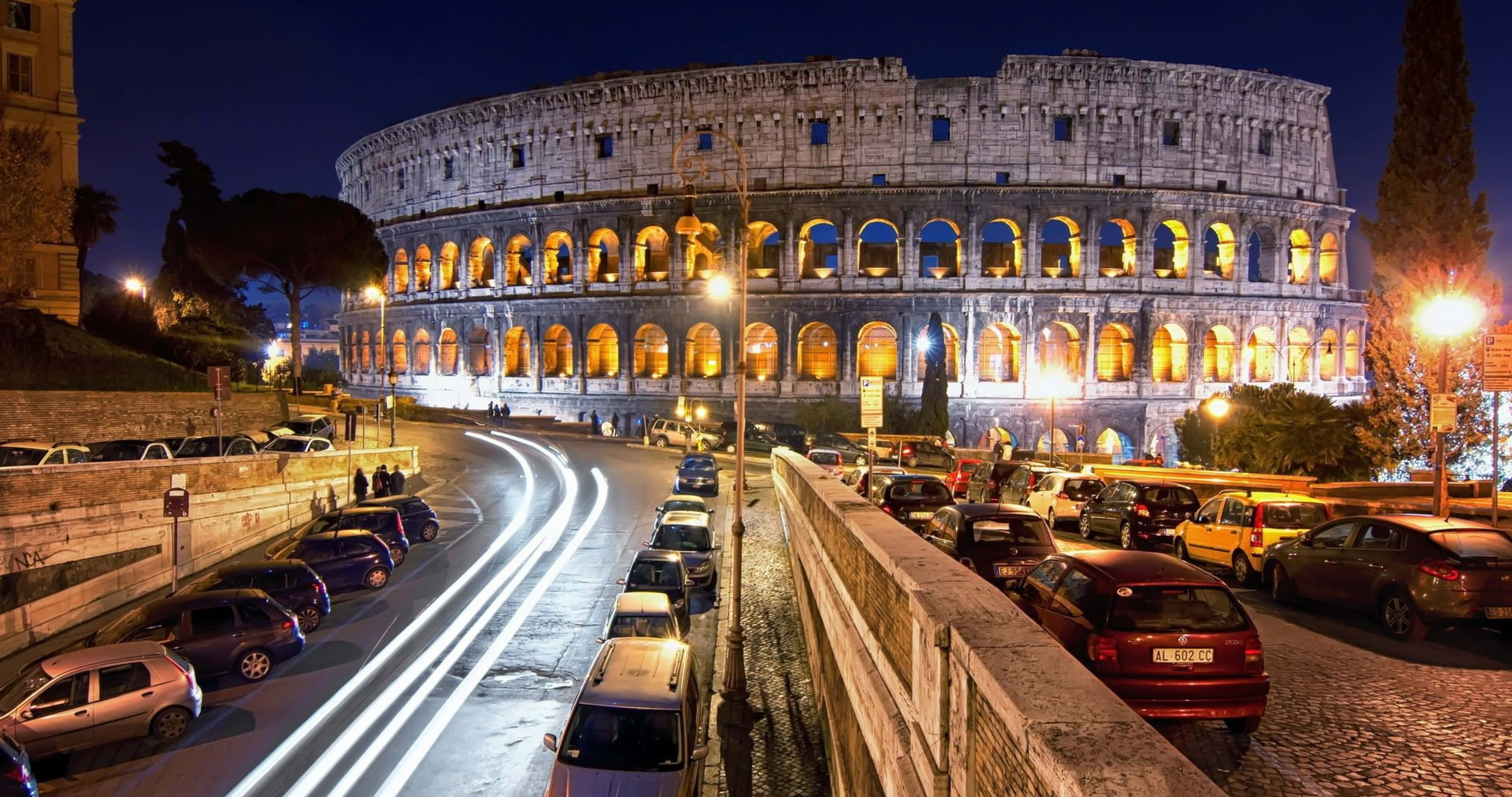 Rome 桌布 壁紙 — 羅馬競技場在夜晚溫暖地發光，而經過的汽車留下的光跡在下方潮濕的道路上劃過。
