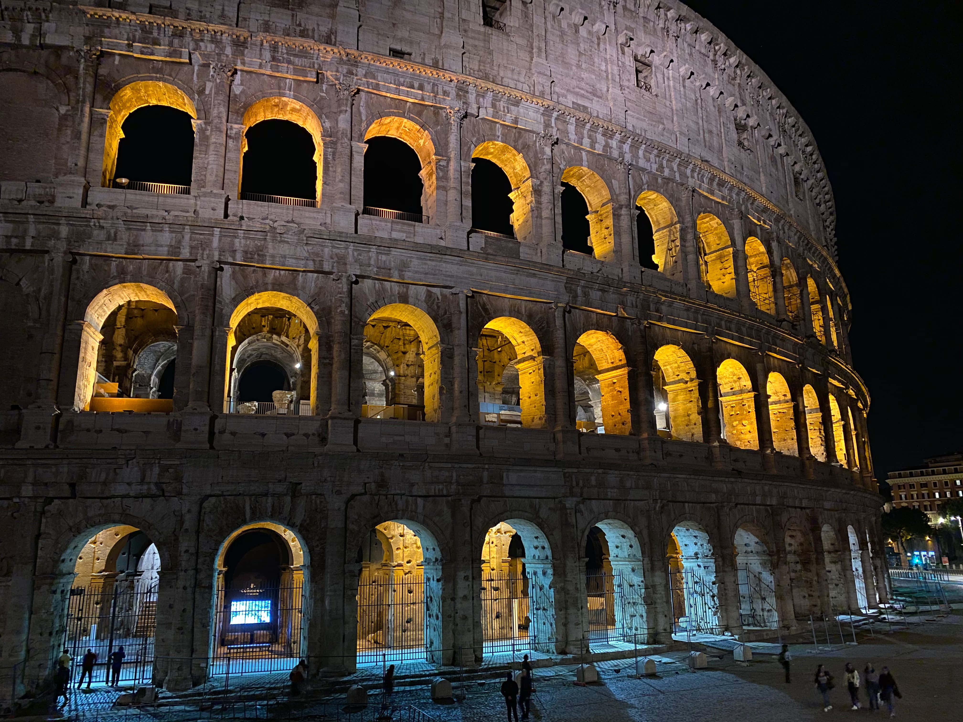 Night Mode 桌布 壁紙 — 羅馬競技場在夜間燈光下溫暖地發光，其古老的石拱門在周圍廣場上投下戲劇性的陰影。
