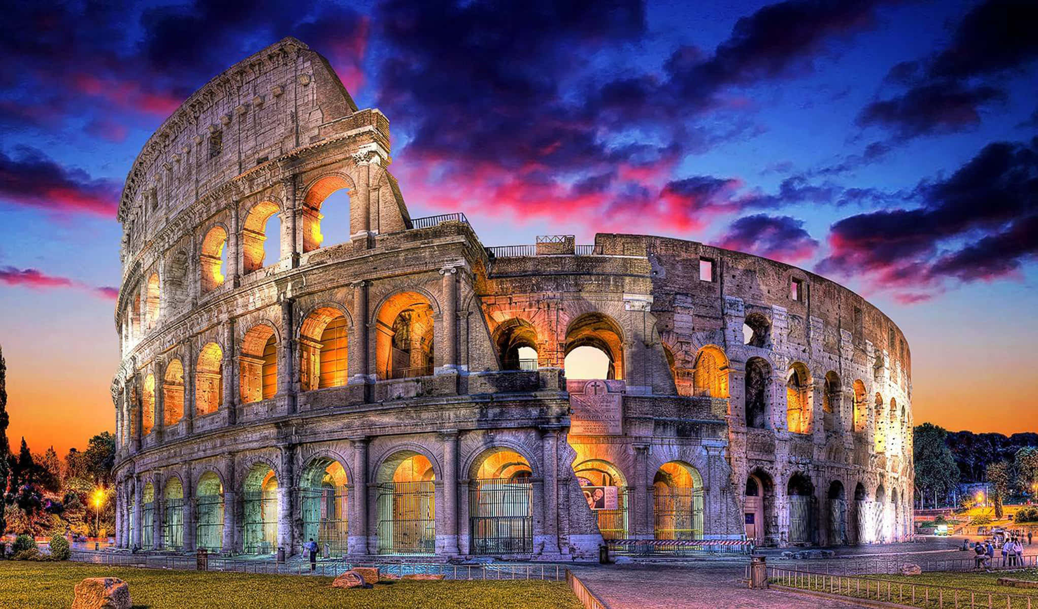 Roman 桌布 壁紙 — 羅馬競技場在戲劇性的黃昏天空下溫暖地發光，天空被火橙色和深紫色的雲彩塗抹。