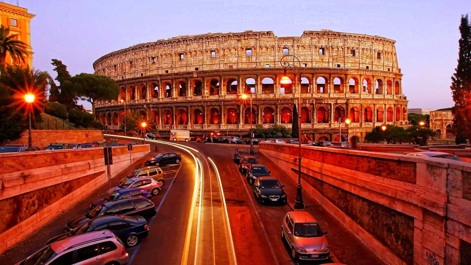Rome 桌布 壁紙 — 羅馬競技場在黃昏時溫暖地發光，下方蜿蜒的道路上車燈劃過。