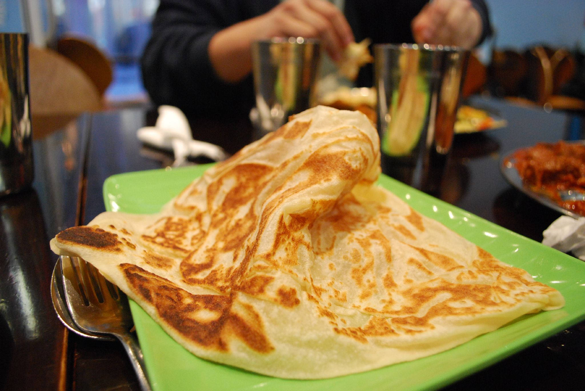 美味Roti Canai 壁紙 — 一個金黃色的折疊印度煎餅放在一個亮綠色的碟子上，旁邊有叉子，在餐廳環境中顯得格外誘人。