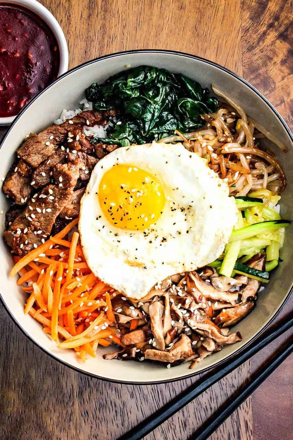 美味多層次的拌飯 壁紙 — 一個充滿活力的韓國石鍋拌飯碗，上面放著太陽蛋、芝麻和色彩繽紛的蔬菜，放在木桌上。