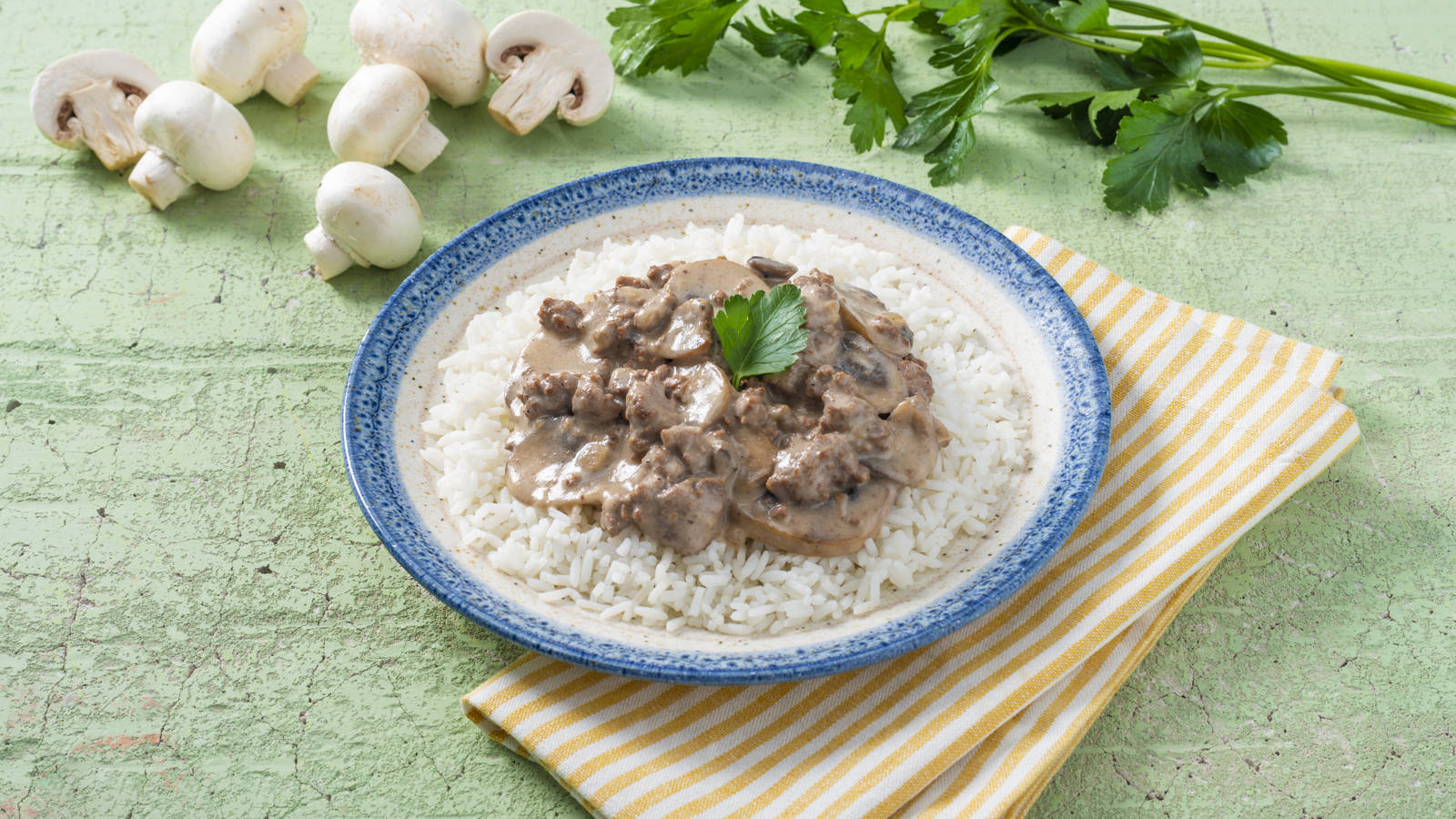 美味牛肉斯特羅甘諾夫配白飯 壁紙 — 一碗奶油蘑菇牛肉燉飯放在白飯上，飾以歐芹，置於條紋餐巾上，背景為有紋理的綠色。