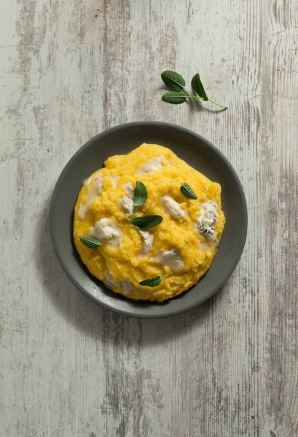 美味玉米粥配義大利餃 壁紙 — 一道鮮黃色的玉米粥菜餚，上面放有奶油般的白色芝士和新鮮的綠色鼠尾草葉，盛在一個質樸的灰色盤子上。