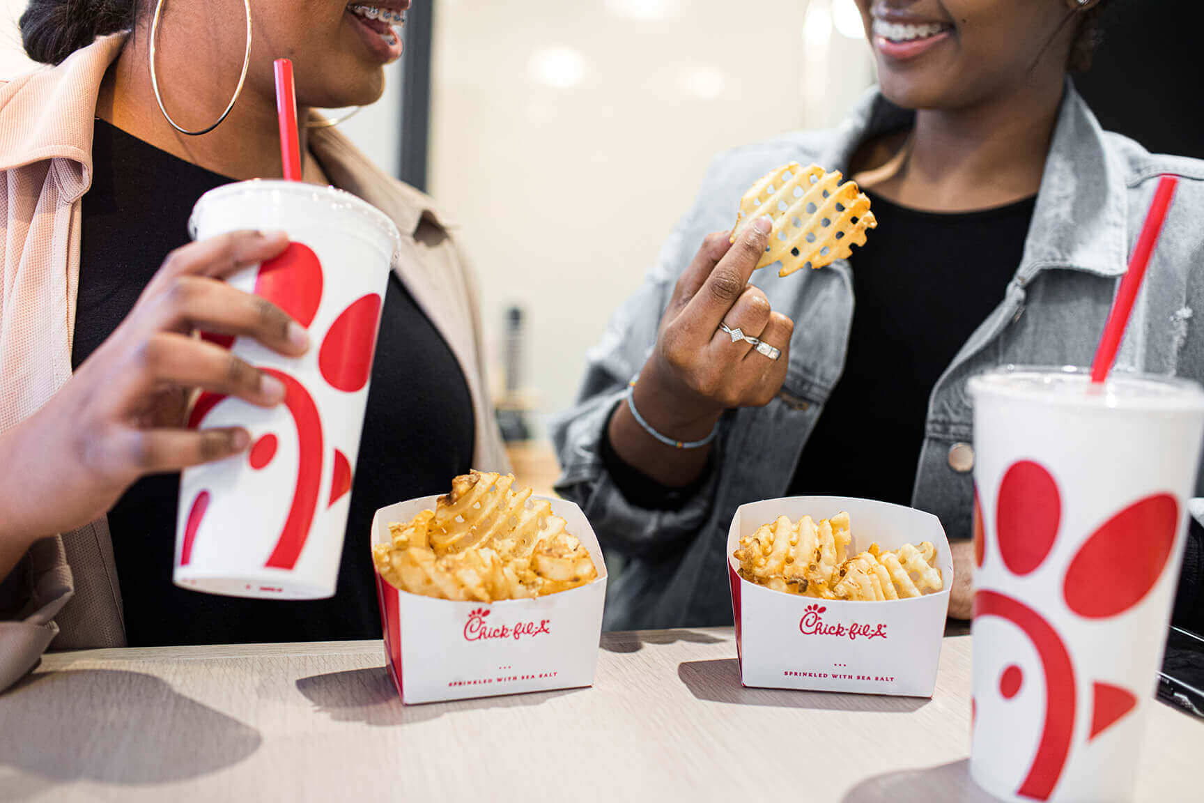 美味的 Chick Fil A 壁紙 — 兩位微笑的女性在休閒餐桌上享用Chick-fil-A的窩夫薯條和飲料。
