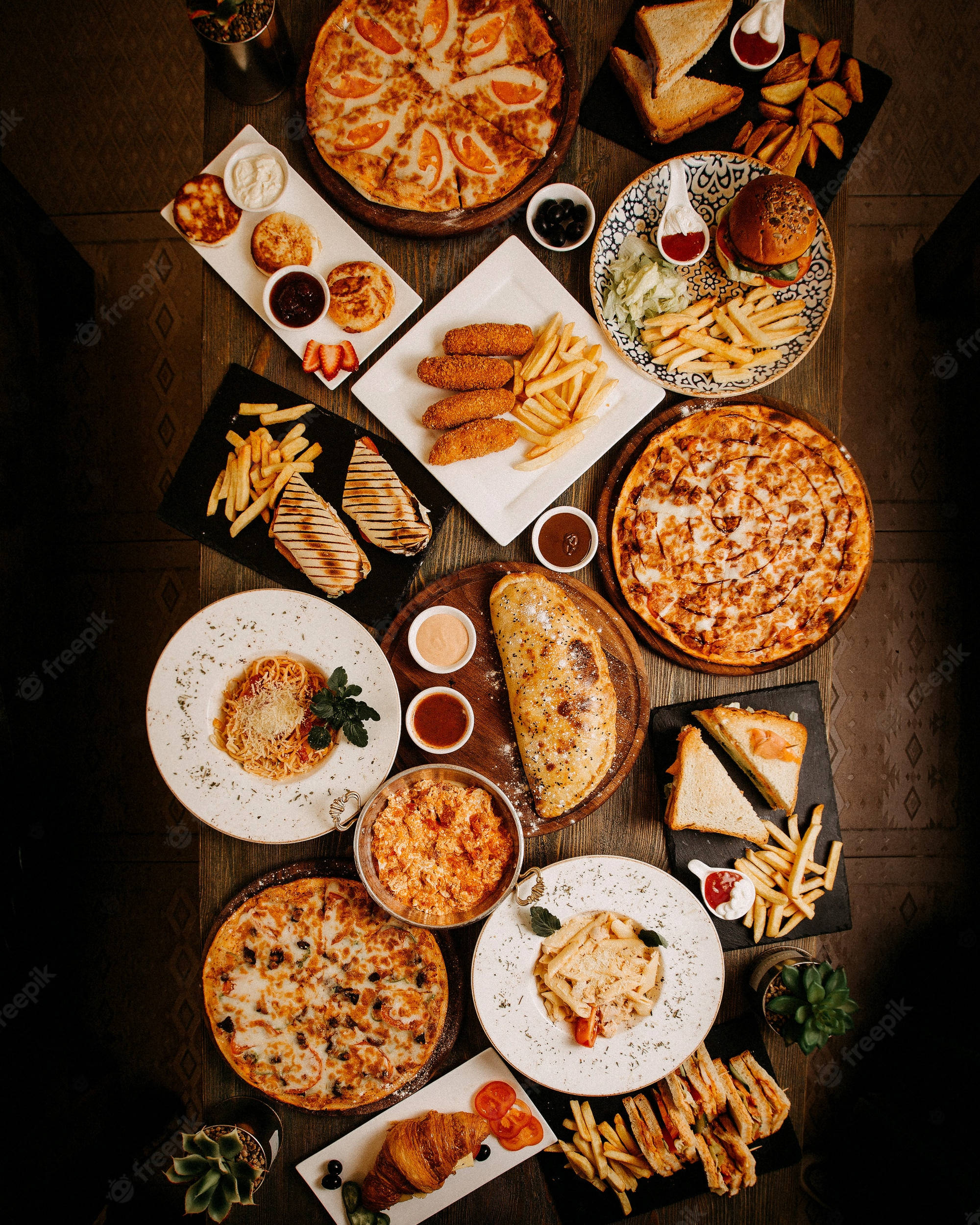美味的披薩及其他美食 壁紙 — 一個豐盛的俯視圖，展示各種快餐菜餚，包括薄餅、漢堡、薯條和意粉，擺放在一張質樸的木桌上。