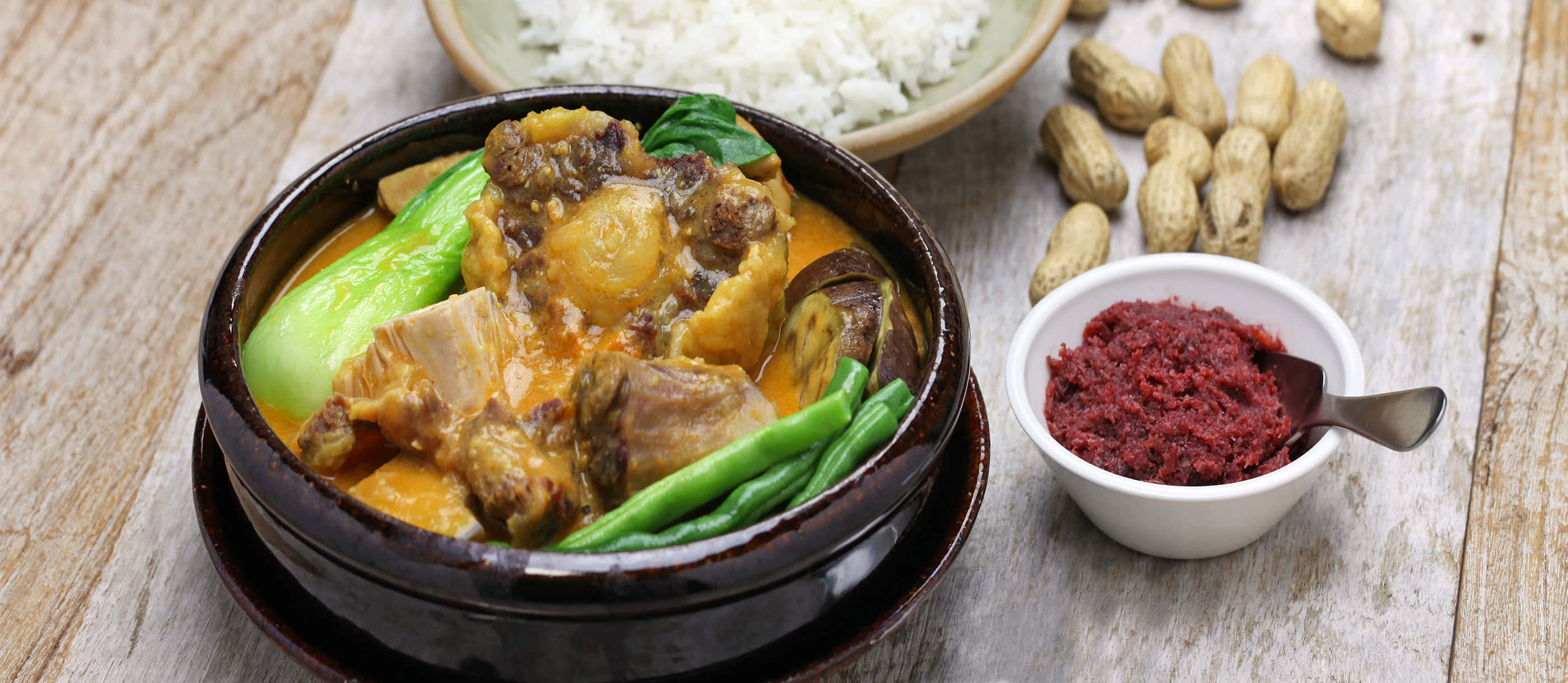 美味的菲律賓燉菜 Kare-Kare 壁紙 — 一碗冒著熱氣的濃郁黃咖喱，配有肉類和綠色蔬菜，旁邊放著白飯、花生和紅辣椒醬，擺在質樸的木桌上。
