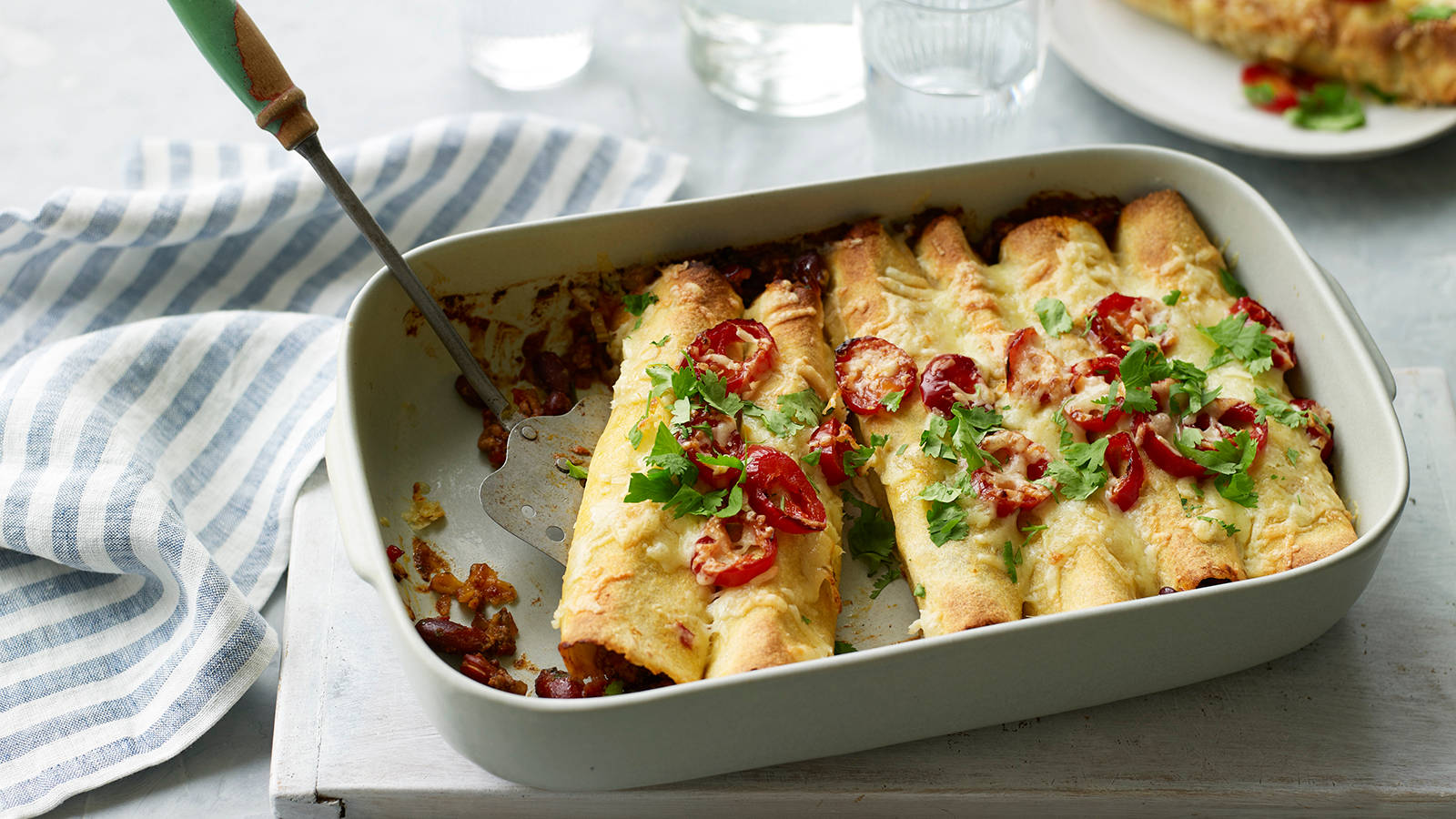 美味起司 enchiladas 卷 壁紙 — 白色烤盤中的金黃色玉米捲餅，上面鋪滿融化芝士、紅椒片和新鮮芫荽。