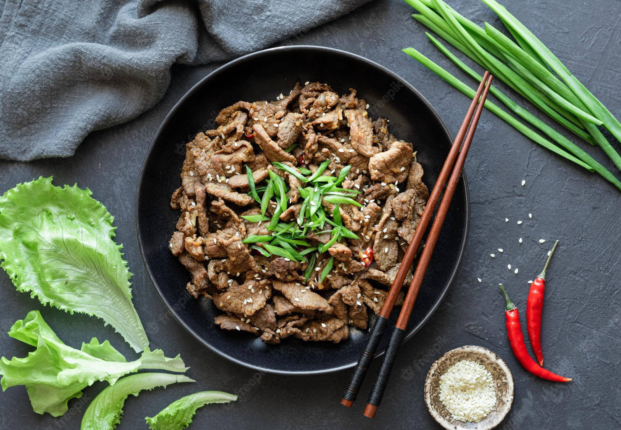 美味韓式醬油牛肉——正宗韓國料理 壁紙 — 一個黑色碗盛載著美味的炒牛肉，上面撒上青蔥、芝麻和紅辣椒，放在深色紋理表面上。