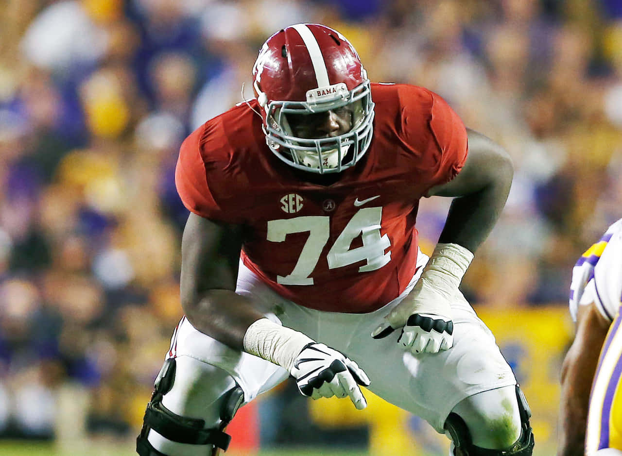 美國 Offensive Tackle 卡姆·羅賓遜三點姿勢 壁紙 — 一名專注的阿拉巴馬州足球員身穿紅色球衣74號，在擠滿模糊球迷的體育館中蹲下。