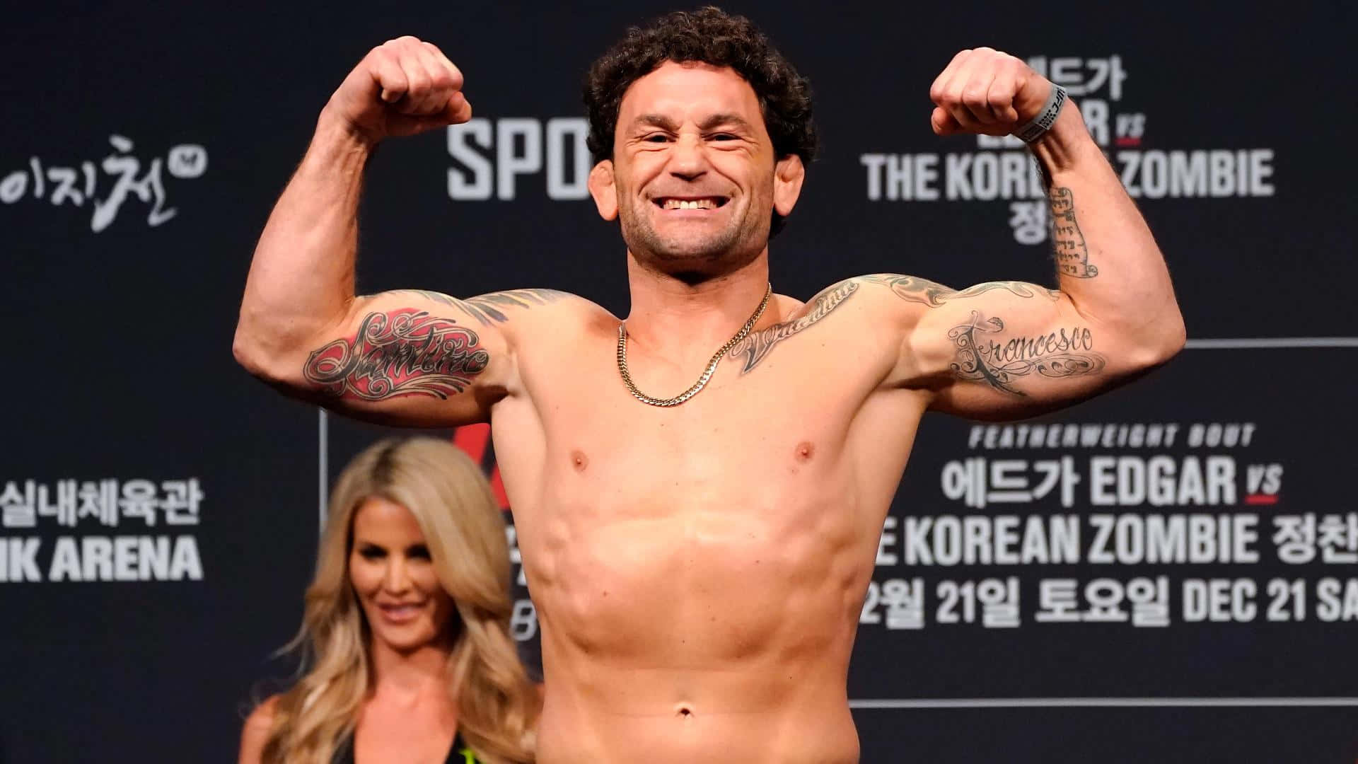 美國 UFC 選手弗蘭基·愛德華稱重儀式 壁紙 — 一位肌肉發達的拳手在稱重活動中彎曲雙臂，露出紋身，微笑著。