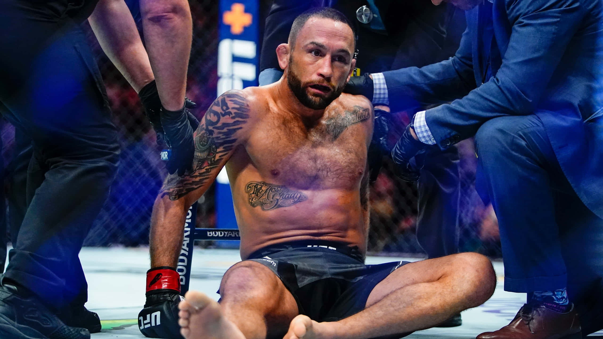 美國 UFC 選手弗蘭基·愛德華被擊倒 壁紙 — 一位紋身的MMA拳手筋疲力竭地坐在八角籠地上，醫療人員在明亮的場館燈光下照顧他。