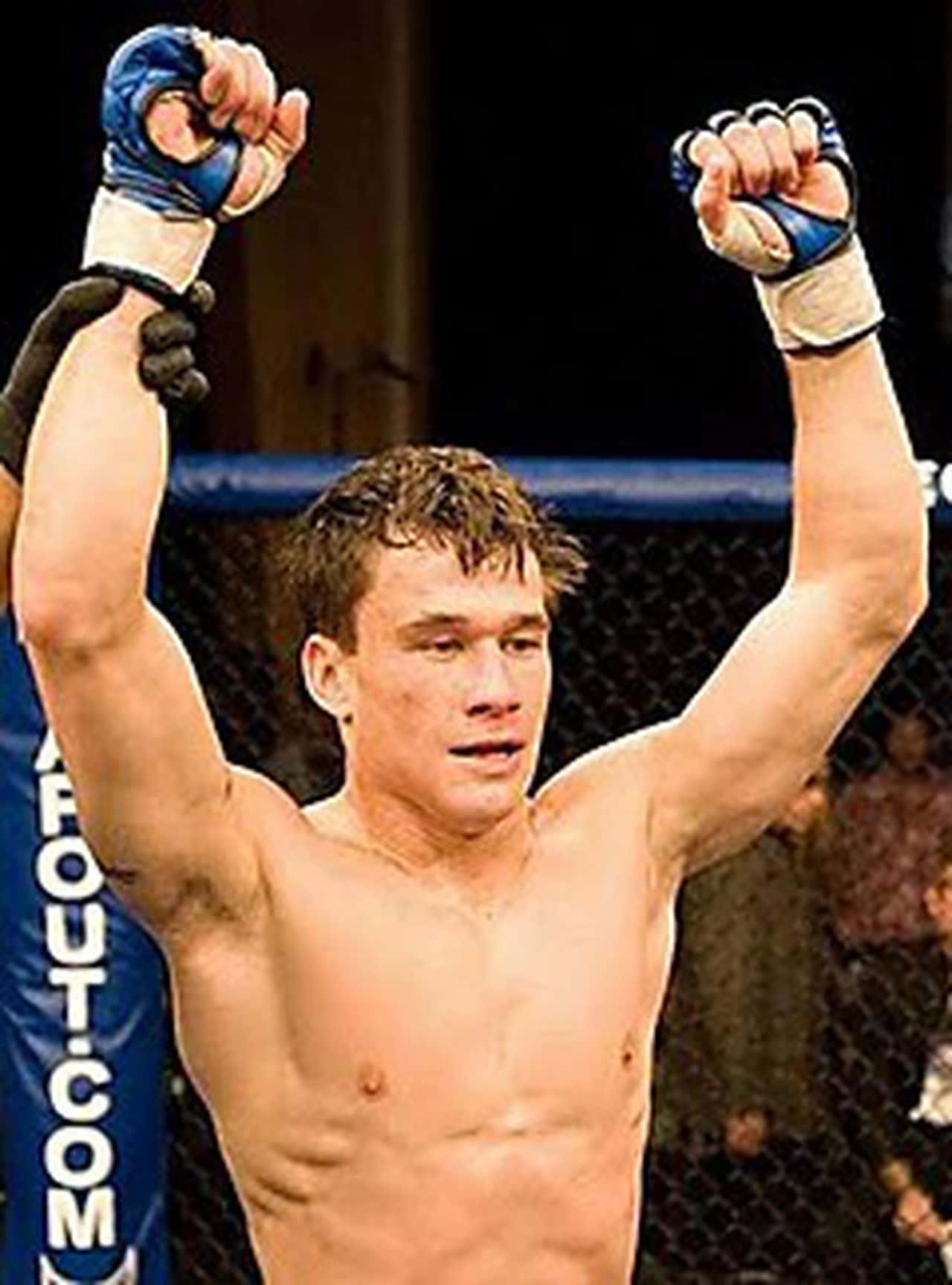 美國MMA戰士布萊恩·鮑爾斯獲勝瞬間 壁紙 — 一位勝利的混合武術家戴著手套的拳頭在昏暗的八角籠內高舉慶祝勝利。